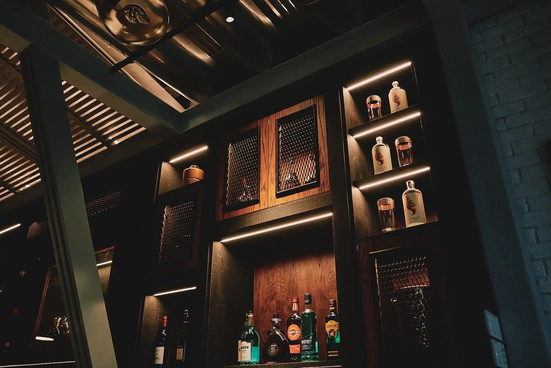 Back bar shelving detail at Prohibition Bar Mandarin Oriental Shenzhen featuring illuminated timber display cabinets with mesh panel doors, curated premium spirit bottles, integrated warm lighting, and exposed ceiling structure reinforcing the speakeasy concept.