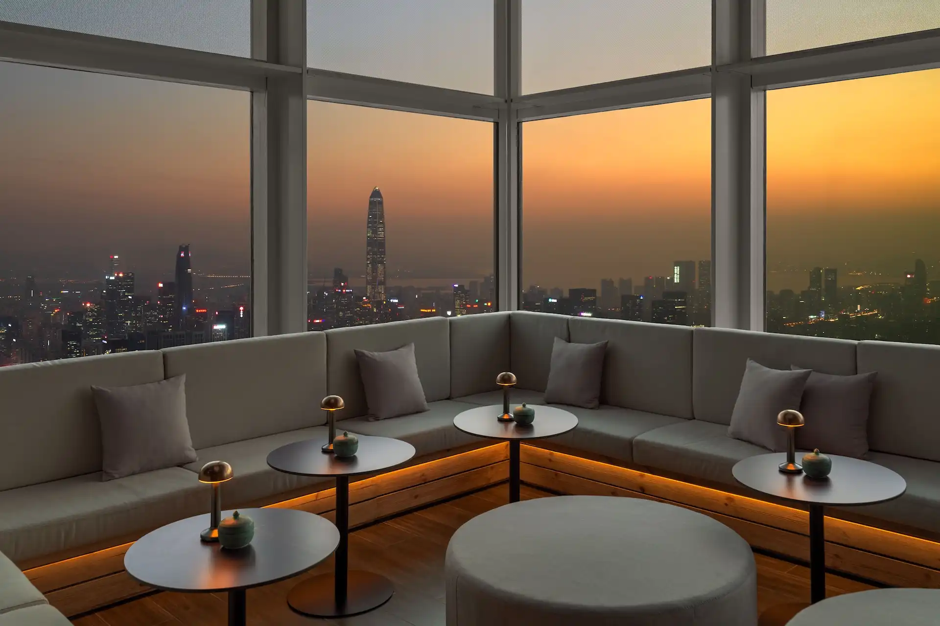 Corner lounge seating at Prohibition Bar Mandarin Oriental Shenzhen with built-in upholstered banquette, circular cocktail tables, integrated warm under-seat lighting, small table lamps, and expansive floor-to-ceiling glazing framing the Shenzhen skyline at sunset.