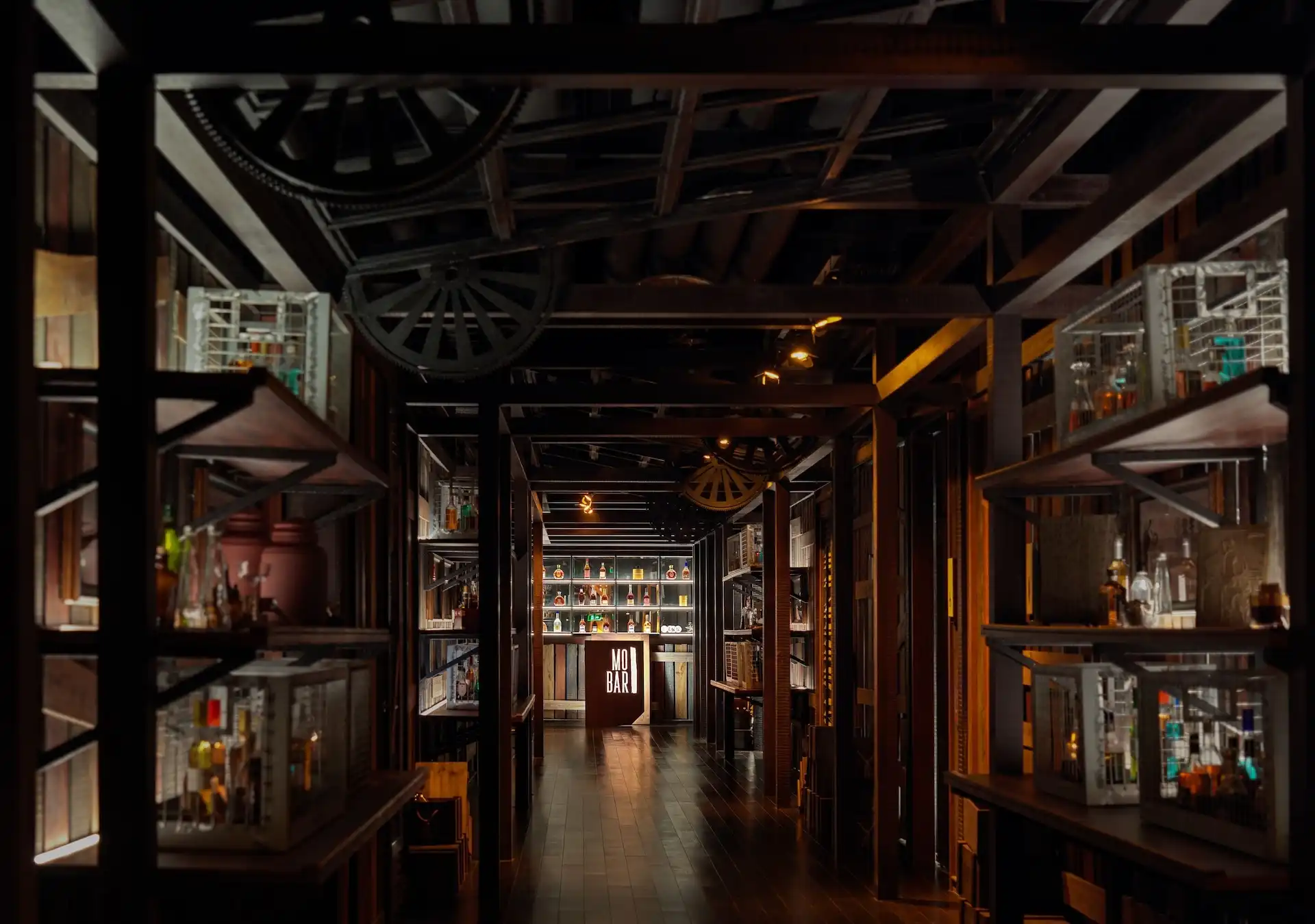 Interior corridor view of Prohibition Bar at Mandarin Oriental Shenzhen featuring a dark timber framework, suspended wagon wheel detailing, illuminated display shelves with liquor bottles, and a central bar counter visible at the end of the passage creating a speakeasy inspired atmosphere.