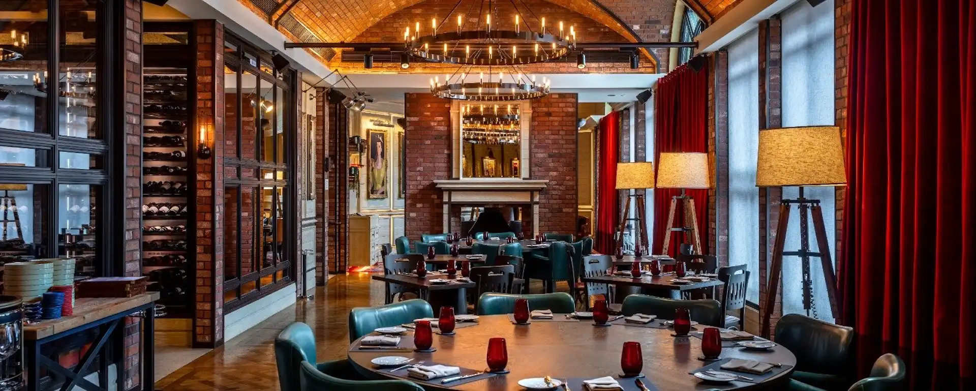 Dining room at Gordon Ramsay Pub & Grill at The Londoner Macao featuring brick vaulted ceilings, circular chandeliers, timber dining tables with red glassware, green leather chairs, wine display shelving, and a fireplace anchoring the space at the far end.