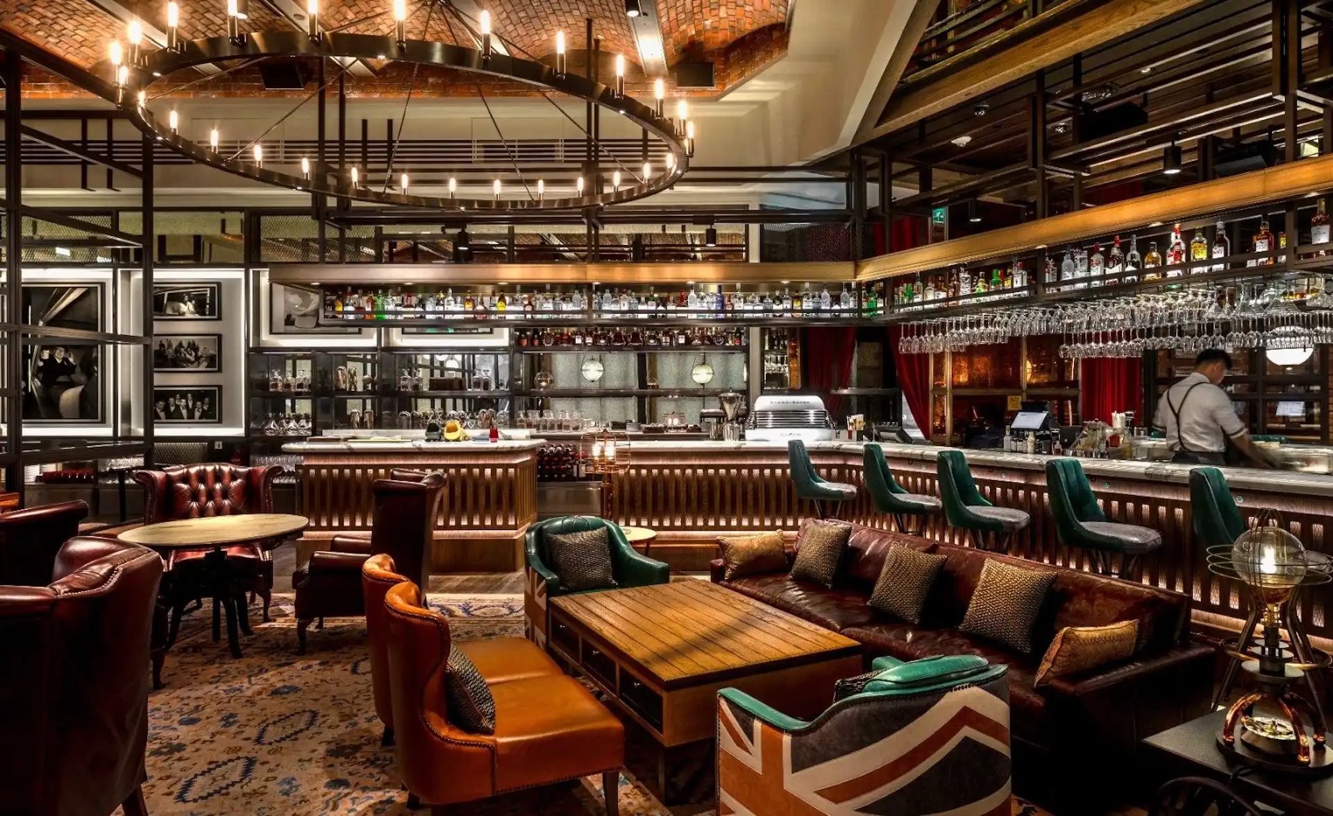 Lounge seating area at Gordon Ramsay Pub & Grill at The Londoner Macao featuring leather sofas, timber coffee tables, patterned carpet, and views toward the illuminated bar counter with suspended bottle racks, hanging glassware, and a large circular chandelier beneath a brick vaulted ceiling.