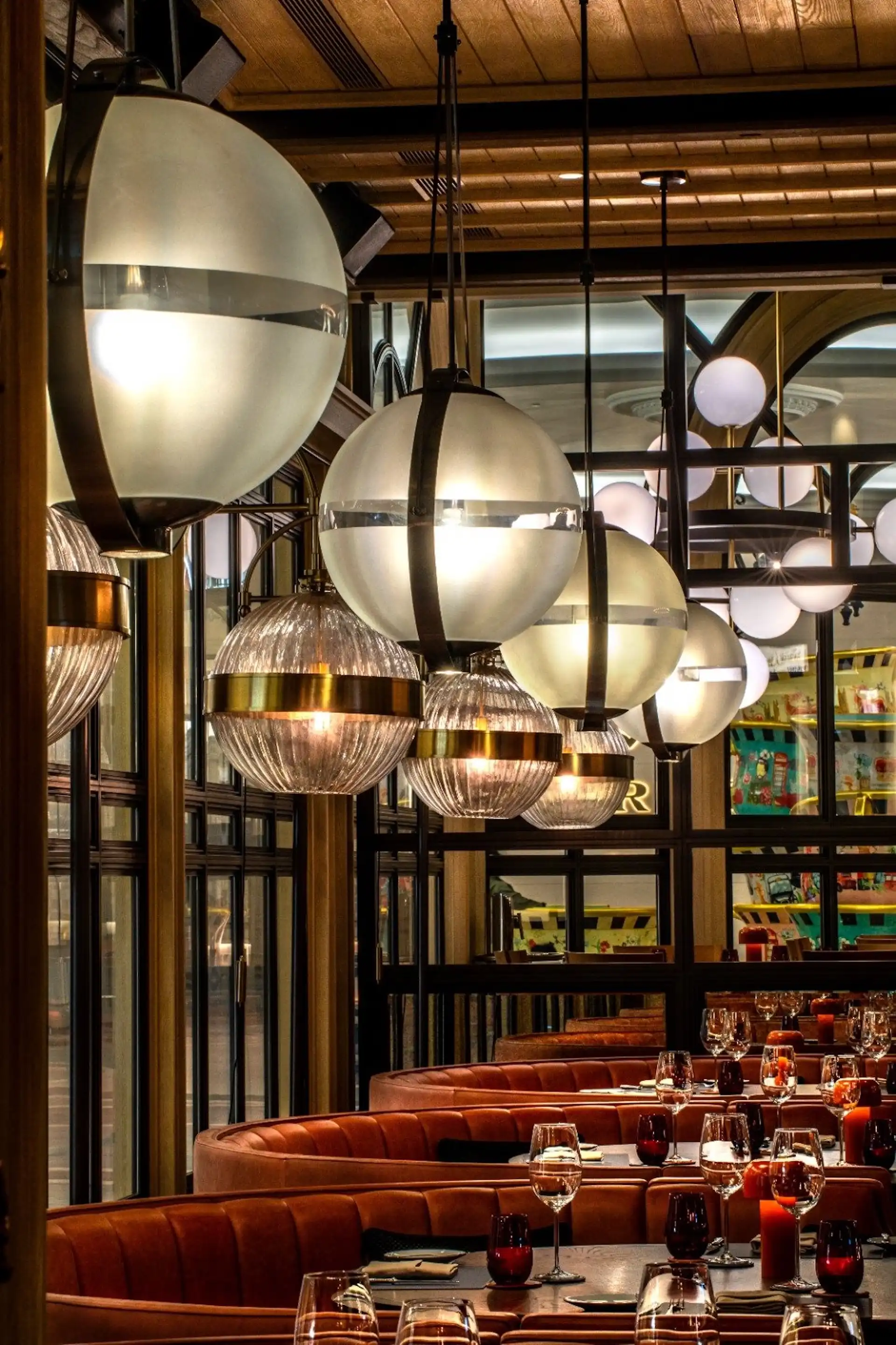 Booth seating area at Gordon Ramsay Pub & Grill at The Londoner Macao featuring curved leather booths, timber dining tables with red glassware, and large spherical pendant lights with brass detailing suspended above the dining space.