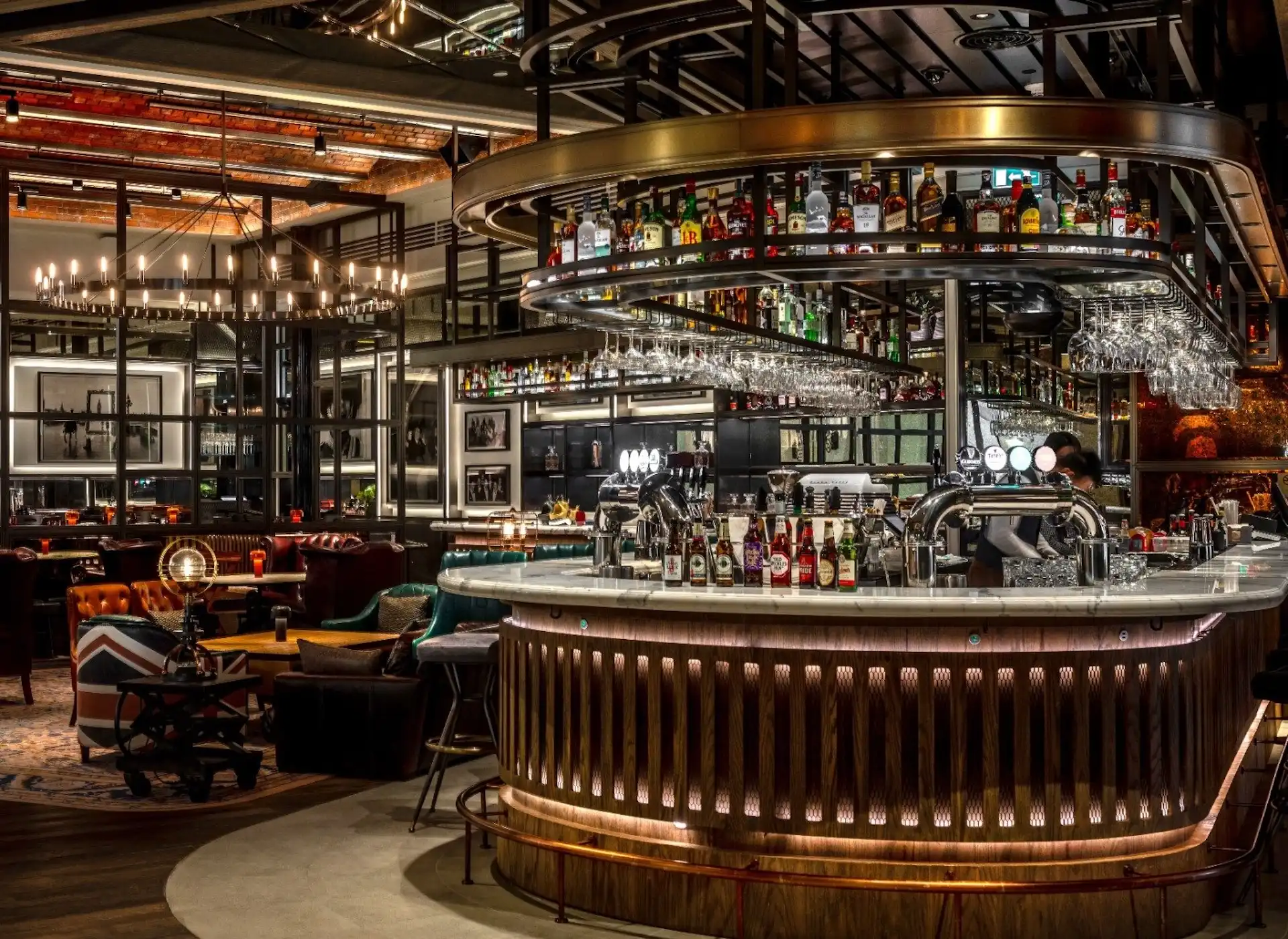Central circular bar at Gordon Ramsay Pub & Grill at The Londoner Macao featuring a marble topped island bar, suspended circular brass bottle rack with hanging glassware, illuminated timber bar front detailing, beer taps, and surrounding lounge seating within a contemporary British pub interior.