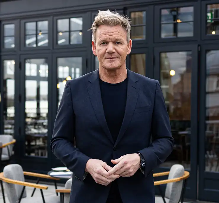 Portrait of chef Gordon Ramsay standing in front of the Gordon Ramsay Pub & Grill restaurant, wearing a dark blazer and shirt, with the restaurant façade visible in the background.