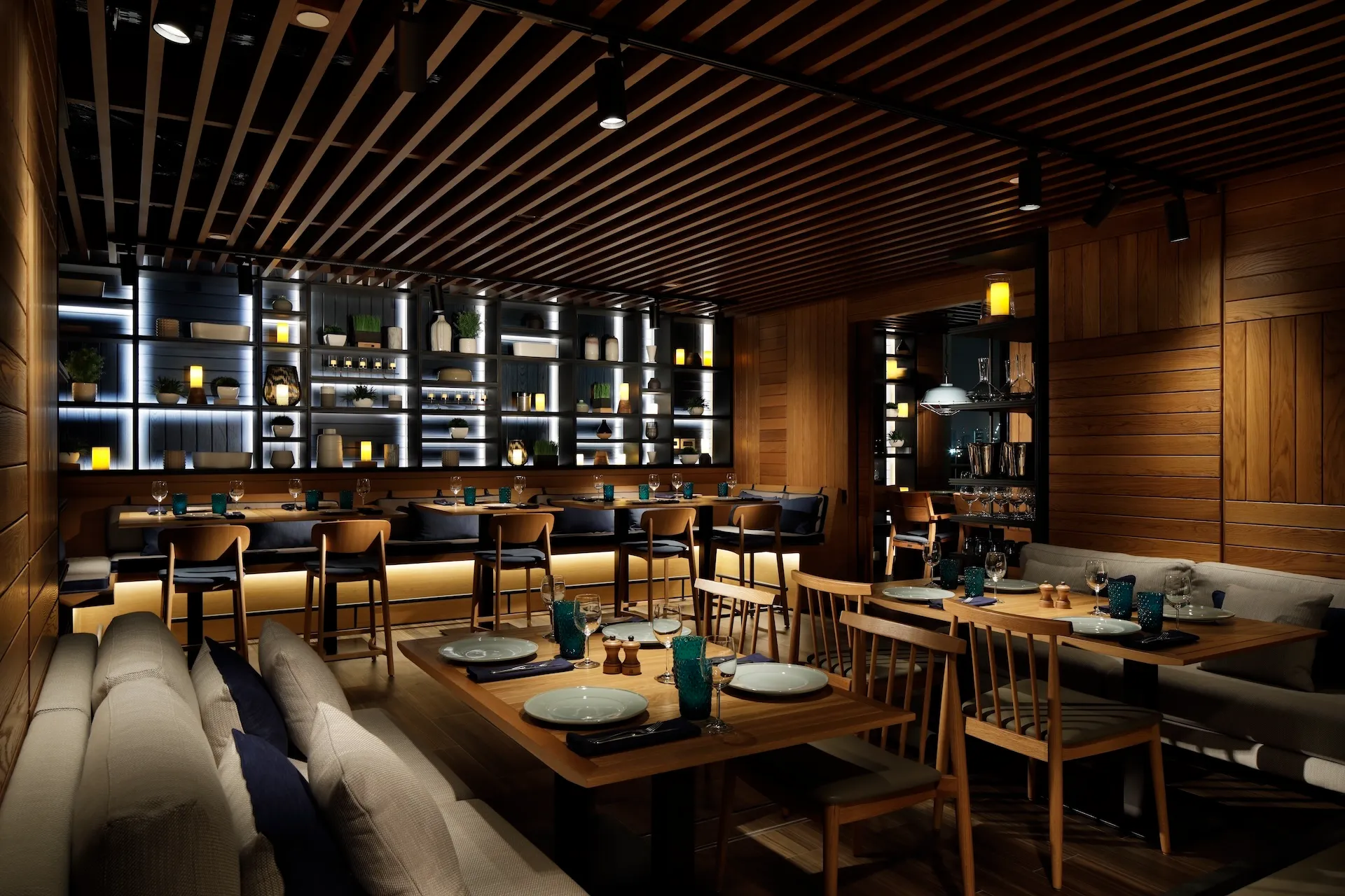 Private dining interior at Tasca by José Avillez at Mandarin Oriental Jumeira Dubai featuring timber tables, upholstered banquette seating, timber slatted ceiling and illuminated shelving display with decorative ceramics and plants.