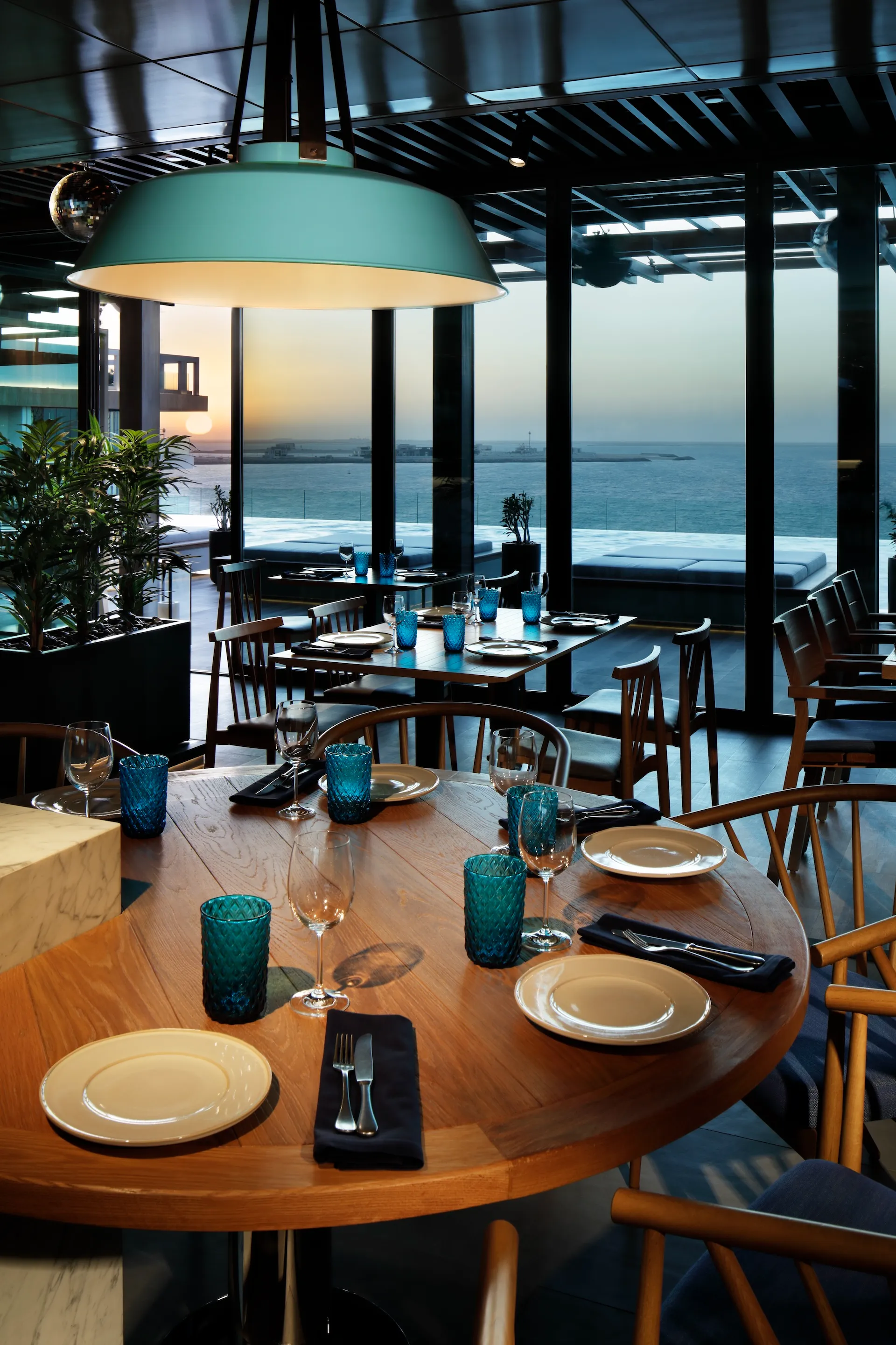 Interior dining space at Tasca by José Avillez at Mandarin Oriental Jumeira Dubai featuring timber tables, wooden chairs, turquoise glassware and floor-to-ceiling glazing overlooking the Arabian Gulf at sunset.