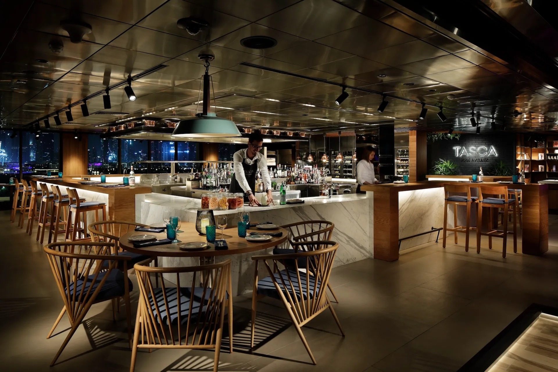 Bar and dining interior at Tasca by José Avillez at Mandarin Oriental Jumeira Dubai featuring marble bar counters, timber seating, turquoise glassware and open bar preparation area with Dubai skyline views at night.