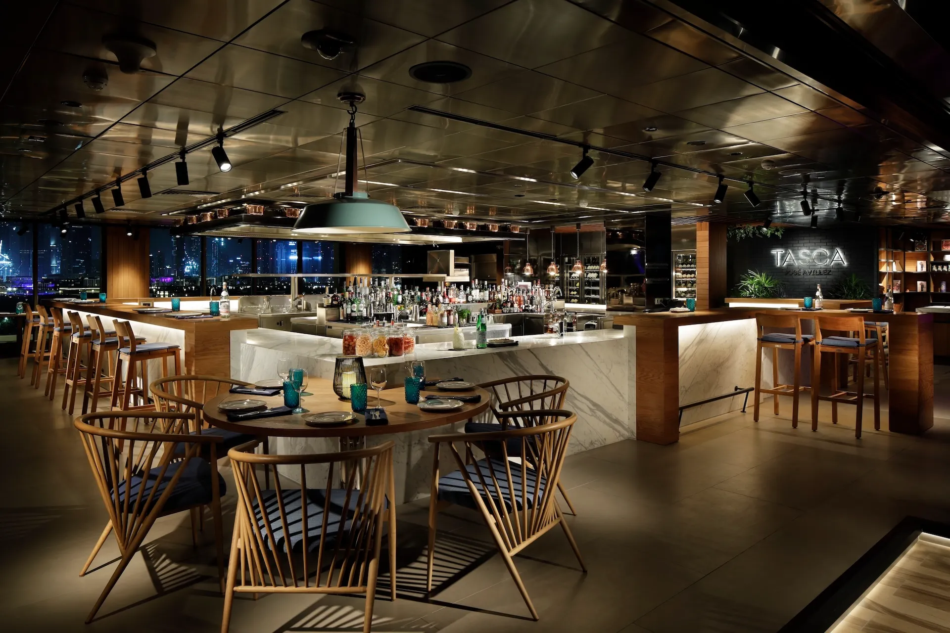 Interior bar and dining space at Tasca by José Avillez at Mandarin Oriental Jumeira Dubai featuring marble bar counters, timber seating, turquoise glassware and open views towards the Dubai skyline at night.