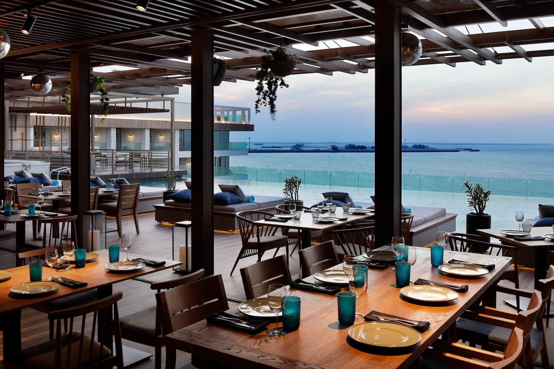 Covered dining terrace at Tasca by José Avillez at Mandarin Oriental Jumeira Dubai with timber dining tables, turquoise glassware, lounge seating and uninterrupted views across the Arabian Gulf.