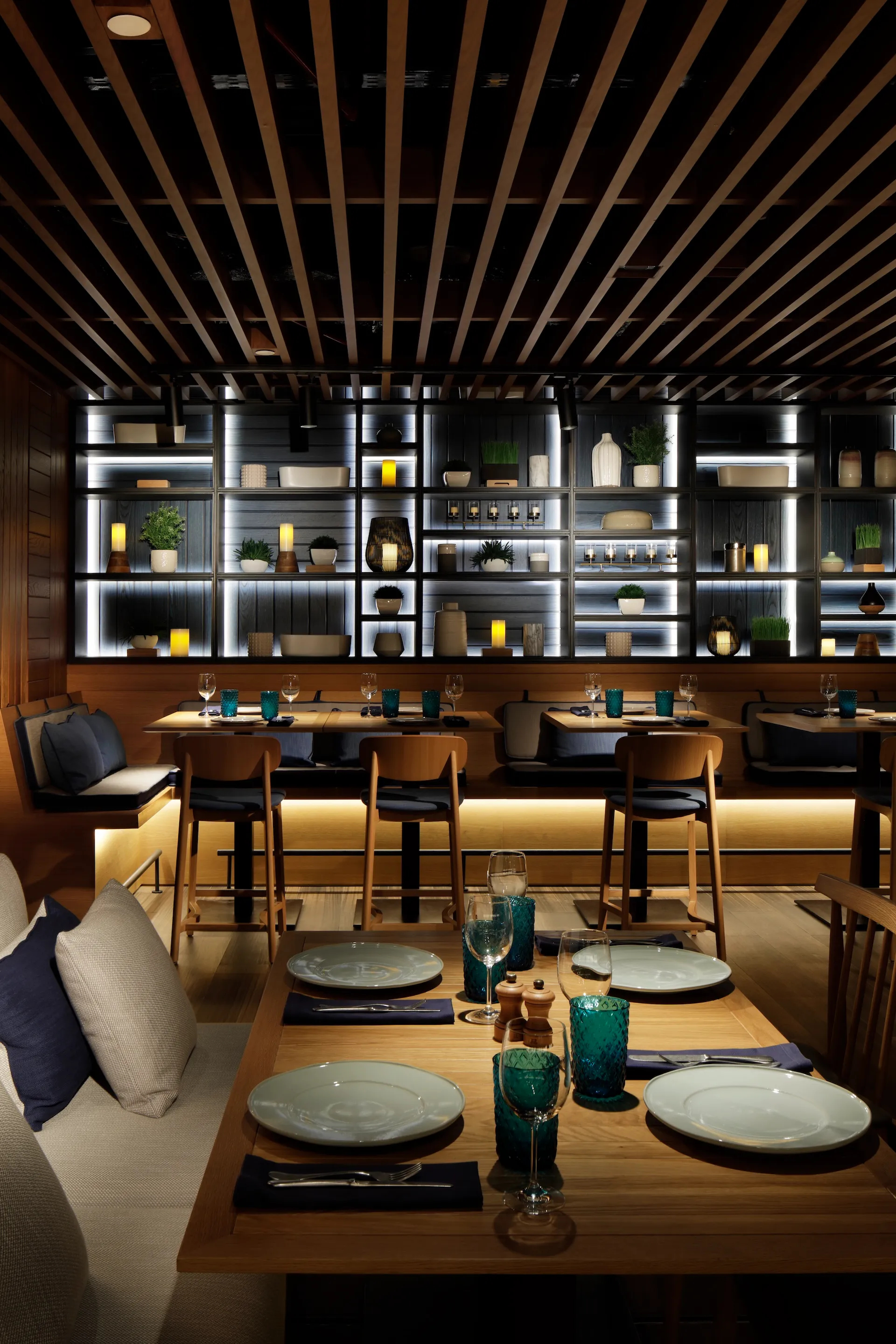 Banquette dining area at Tasca by José Avillez at Mandarin Oriental Jumeira Dubai with timber tables, turquoise glassware, upholstered seating and illuminated shelving displaying ceramics, plants and decorative objects.