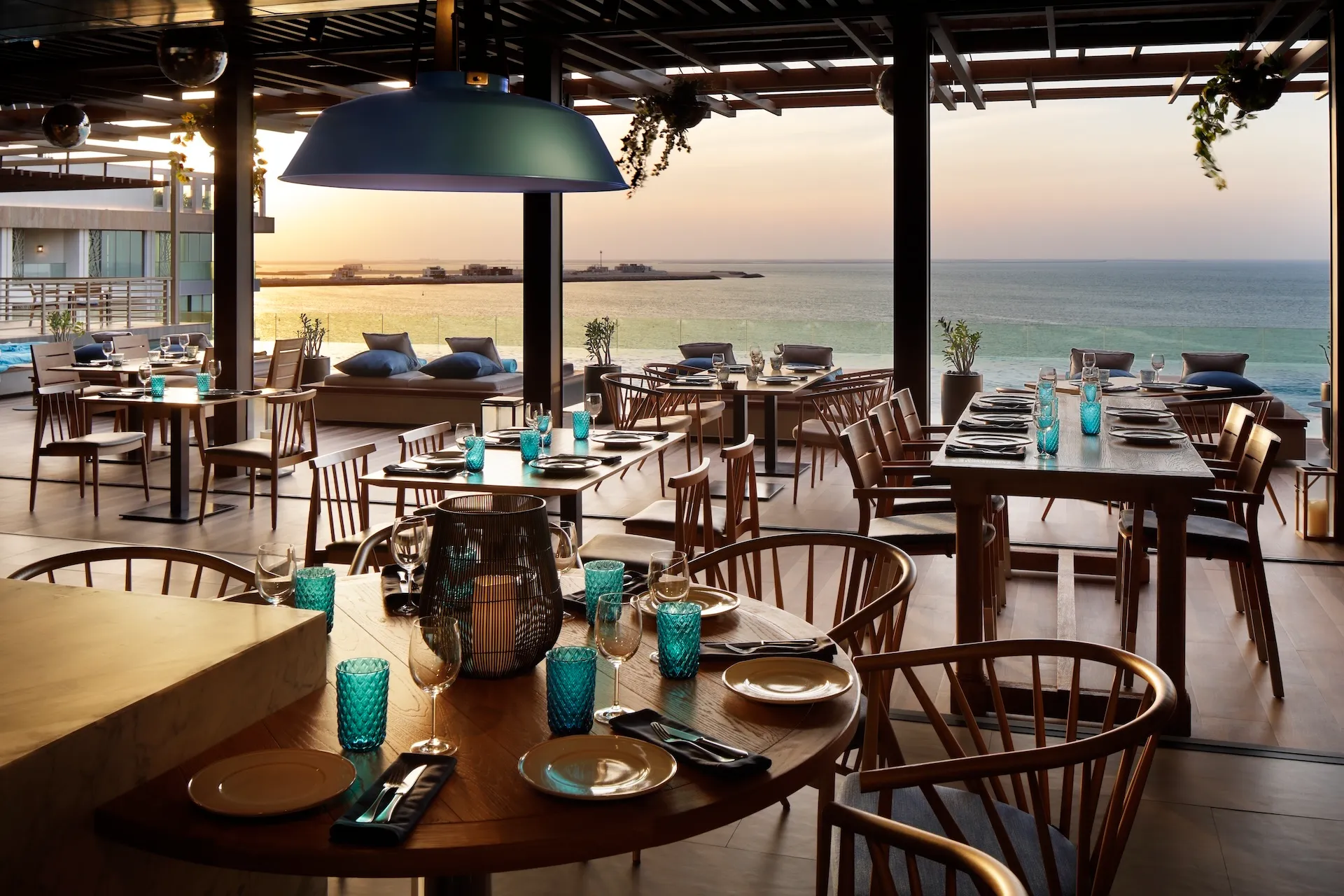 Dining terrace at Tasca by José Avillez at Mandarin Oriental Jumeira Dubai featuring timber tables, wooden chairs, turquoise glassware, and open views across the Arabian Gulf at sunset.