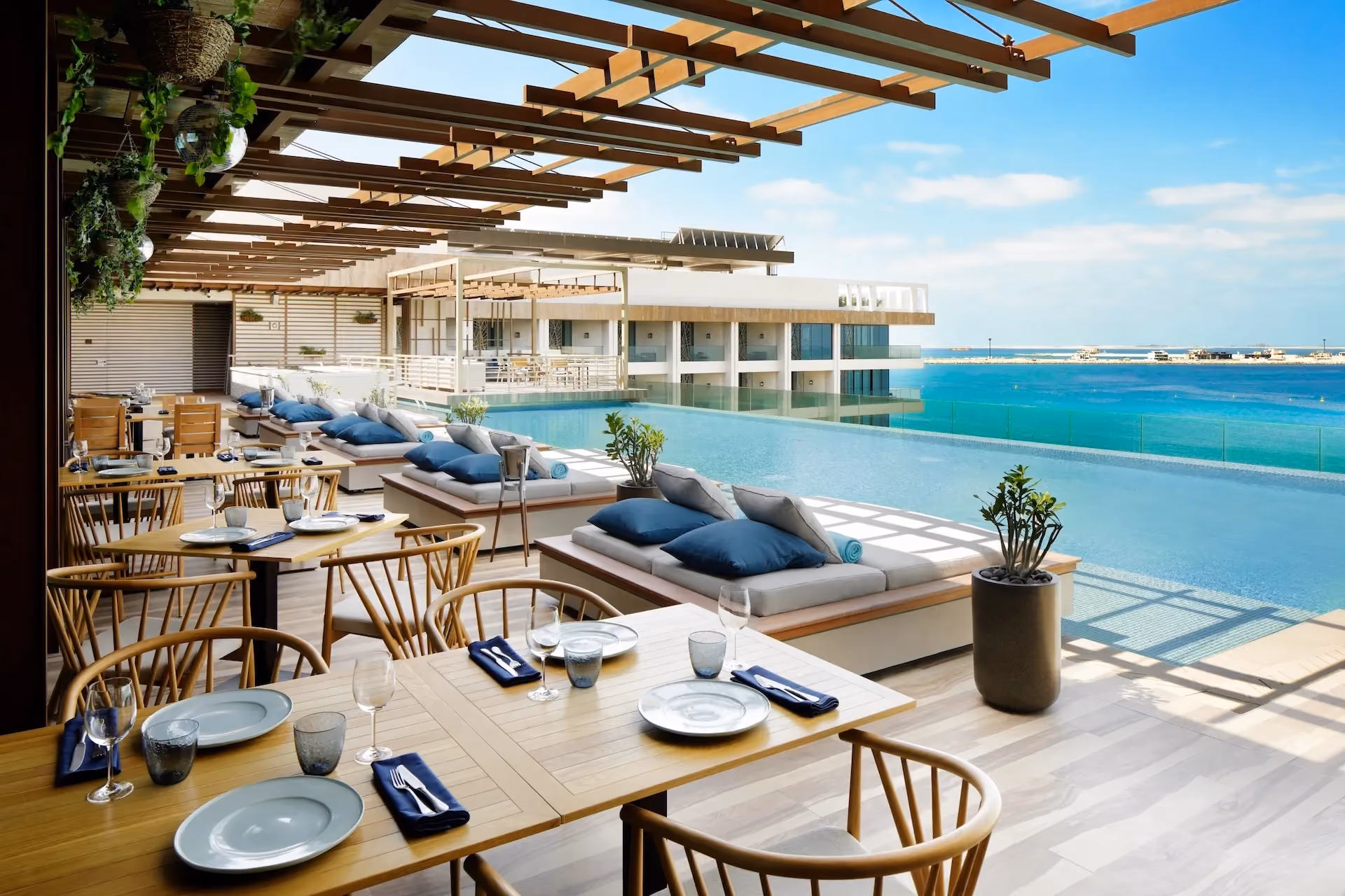 Outdoor terrace at Tasca by José Avillez at Mandarin Oriental Jumeira Dubai featuring timber dining tables, woven chairs, daybeds with blue cushions, a rooftop infinity pool, and views over the Arabian Gulf.