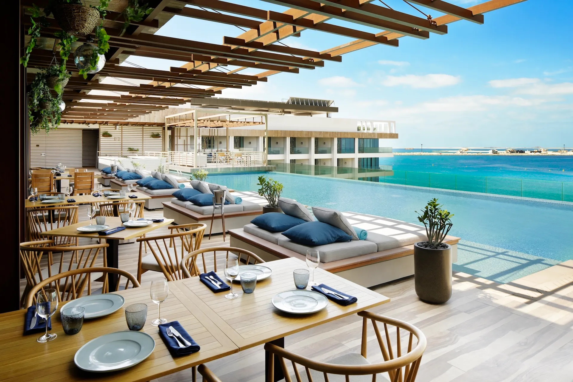 Outdoor terrace at Tasca by José Avillez at Mandarin Oriental Jumeira Dubai featuring timber dining tables, woven chairs, daybeds with blue cushions, a rooftop infinity pool, and views over the Arabian Gulf.