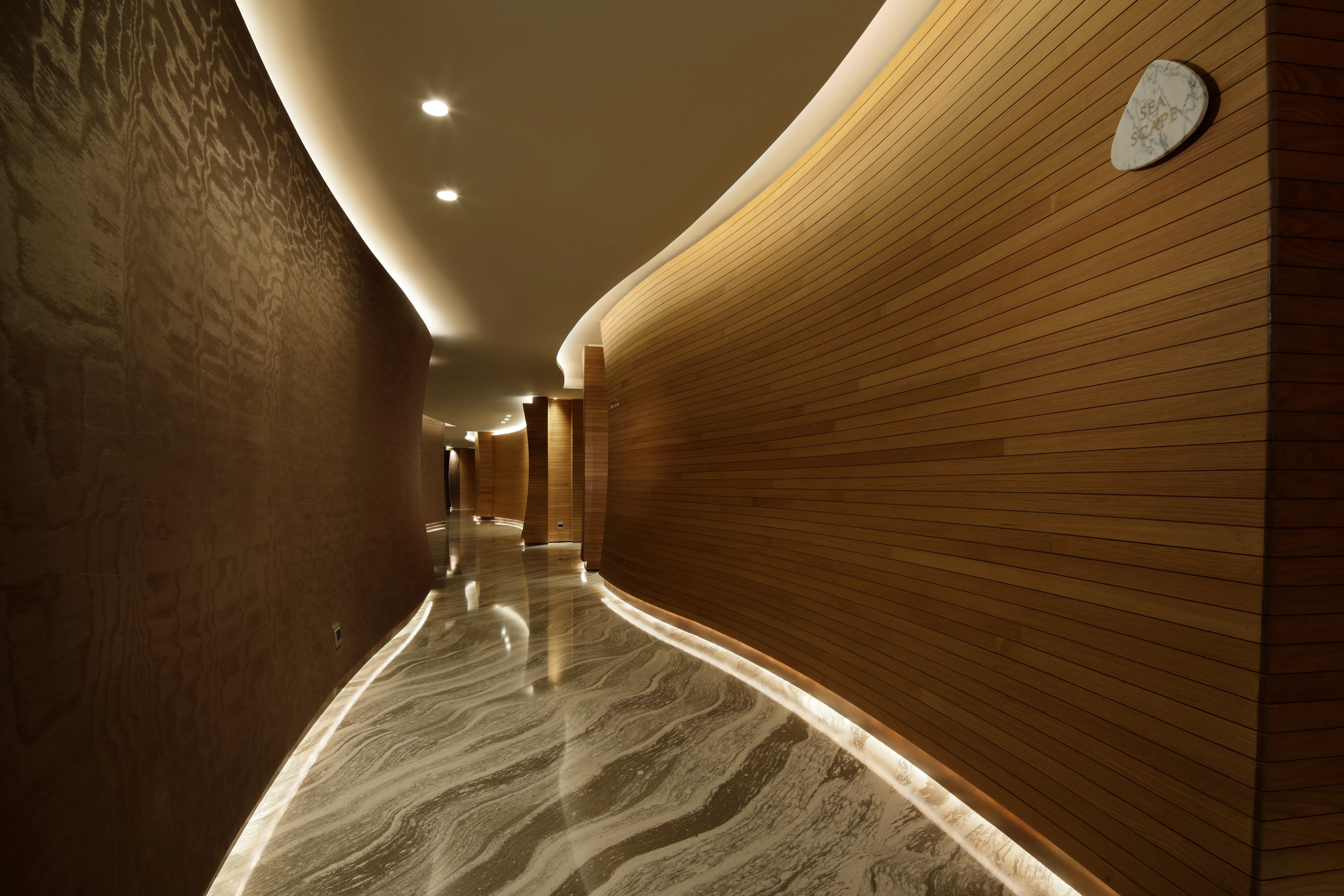 Curved corridor at The Spa, Mandarin Oriental Jumeira Dubai, lined with timber slatting, with cove lighting overhead and uplighting along the marble floor