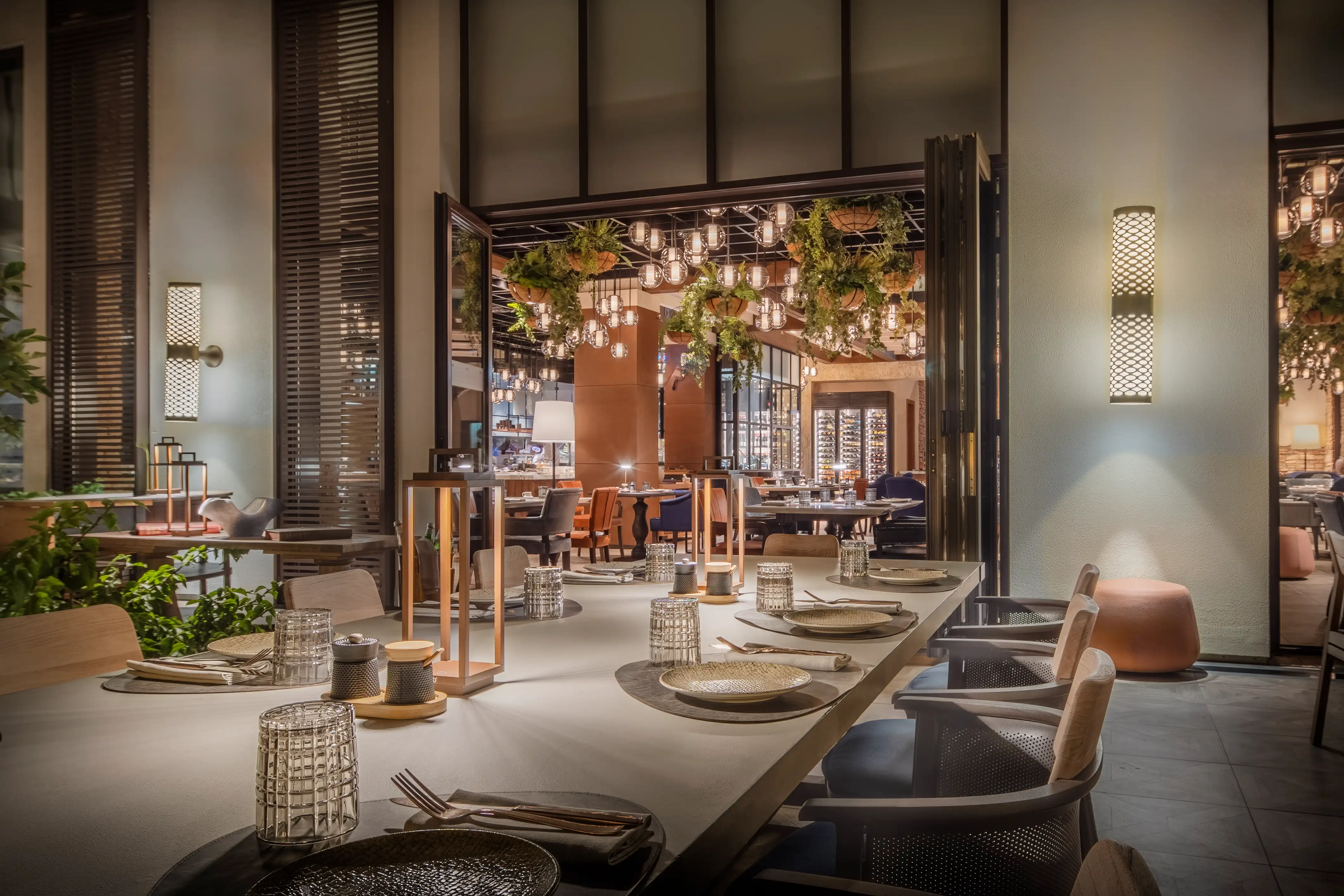 Long dining table beside timber shutters and wall sconces, looking into a room hung with globe pendants, at Essenza, Mandarin Oriental, Muscat