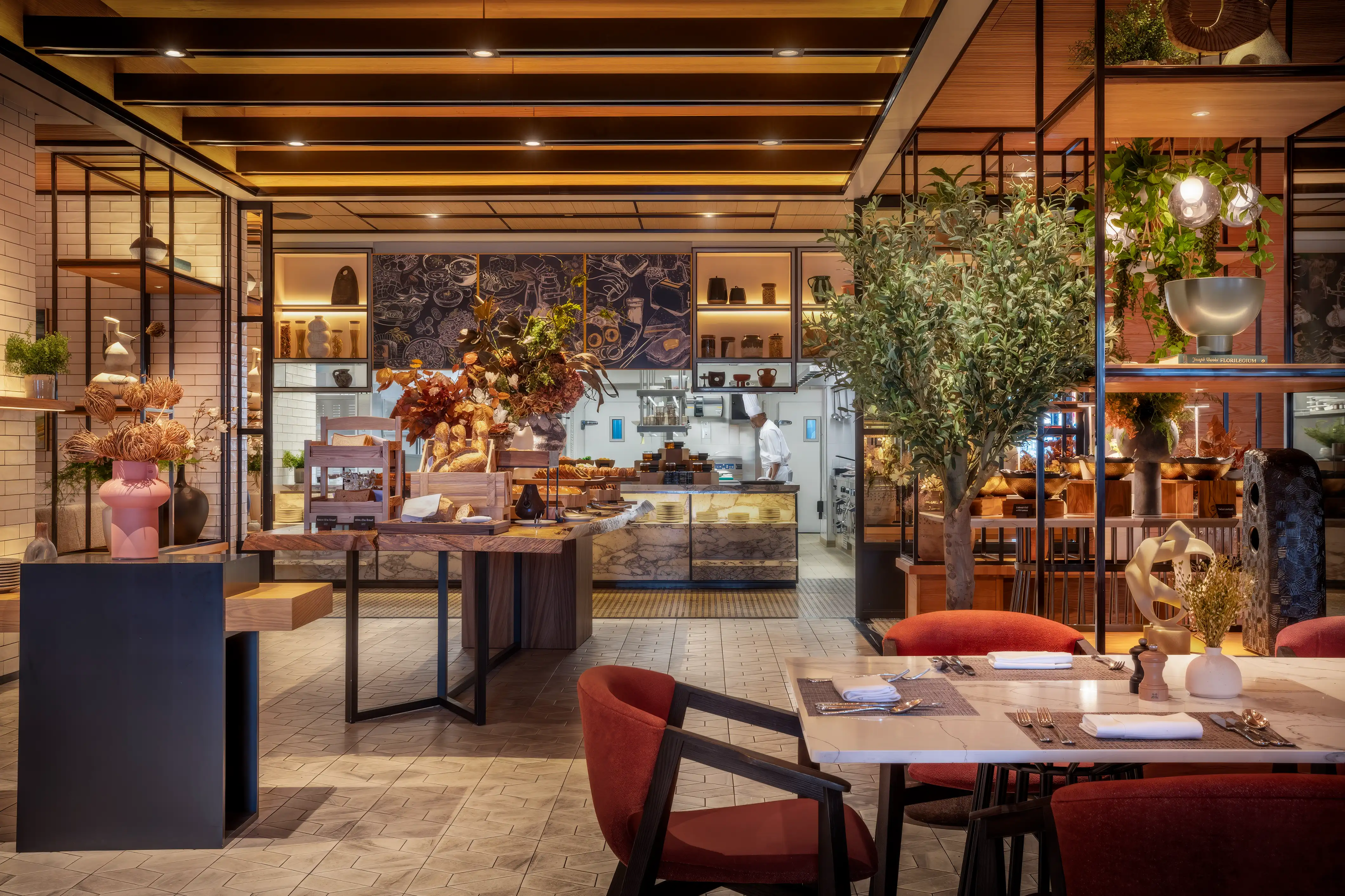 Backlit timber ceiling with dark beams over red dining chairs, a potted tree and the open kitchen at Rawya, Mandarin Oriental, Muscat