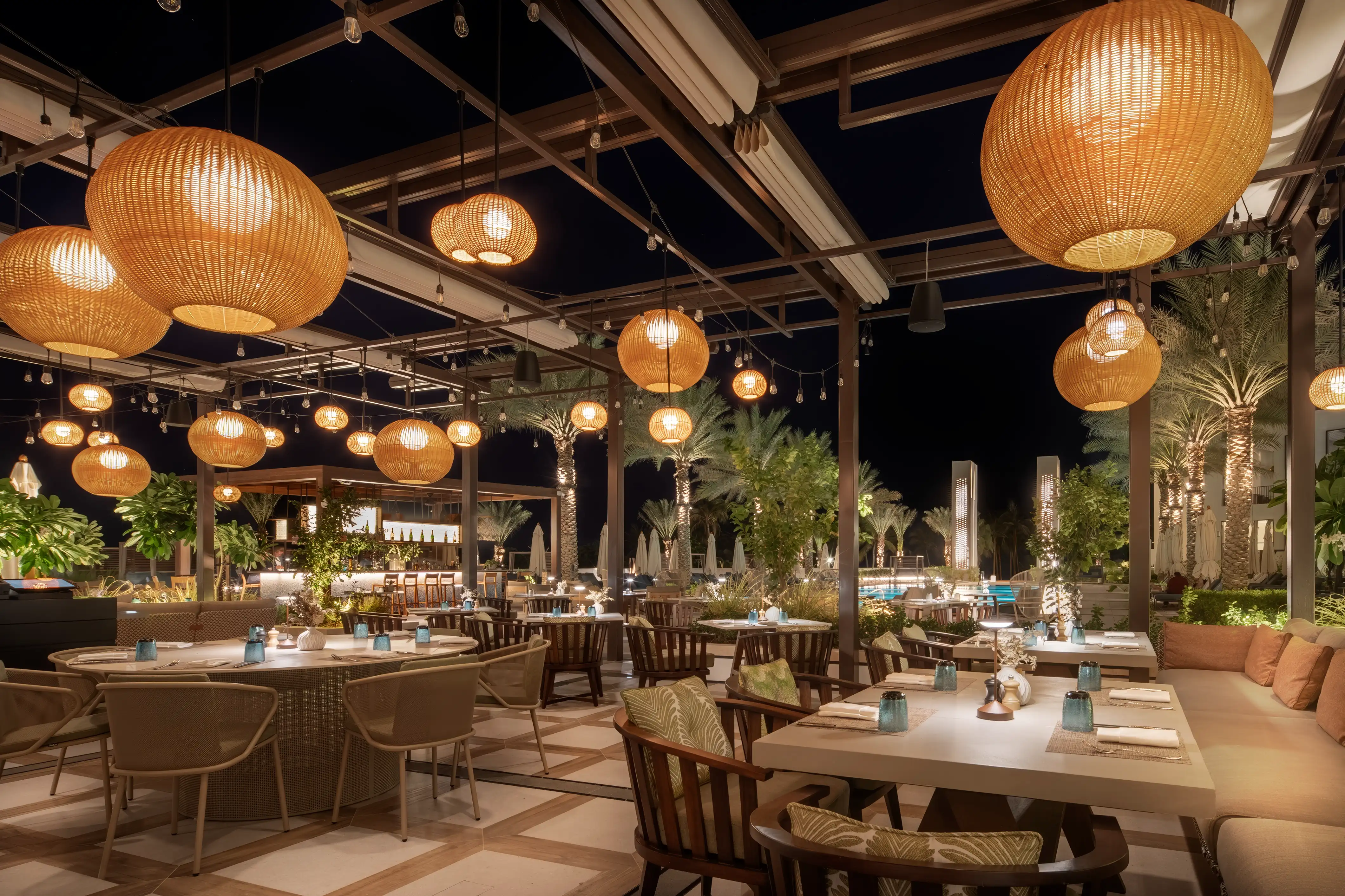 Garden terrace at night with oversized woven pendants over timber dining chairs and palms beyond at Rawya, Mandarin Oriental, Muscat
