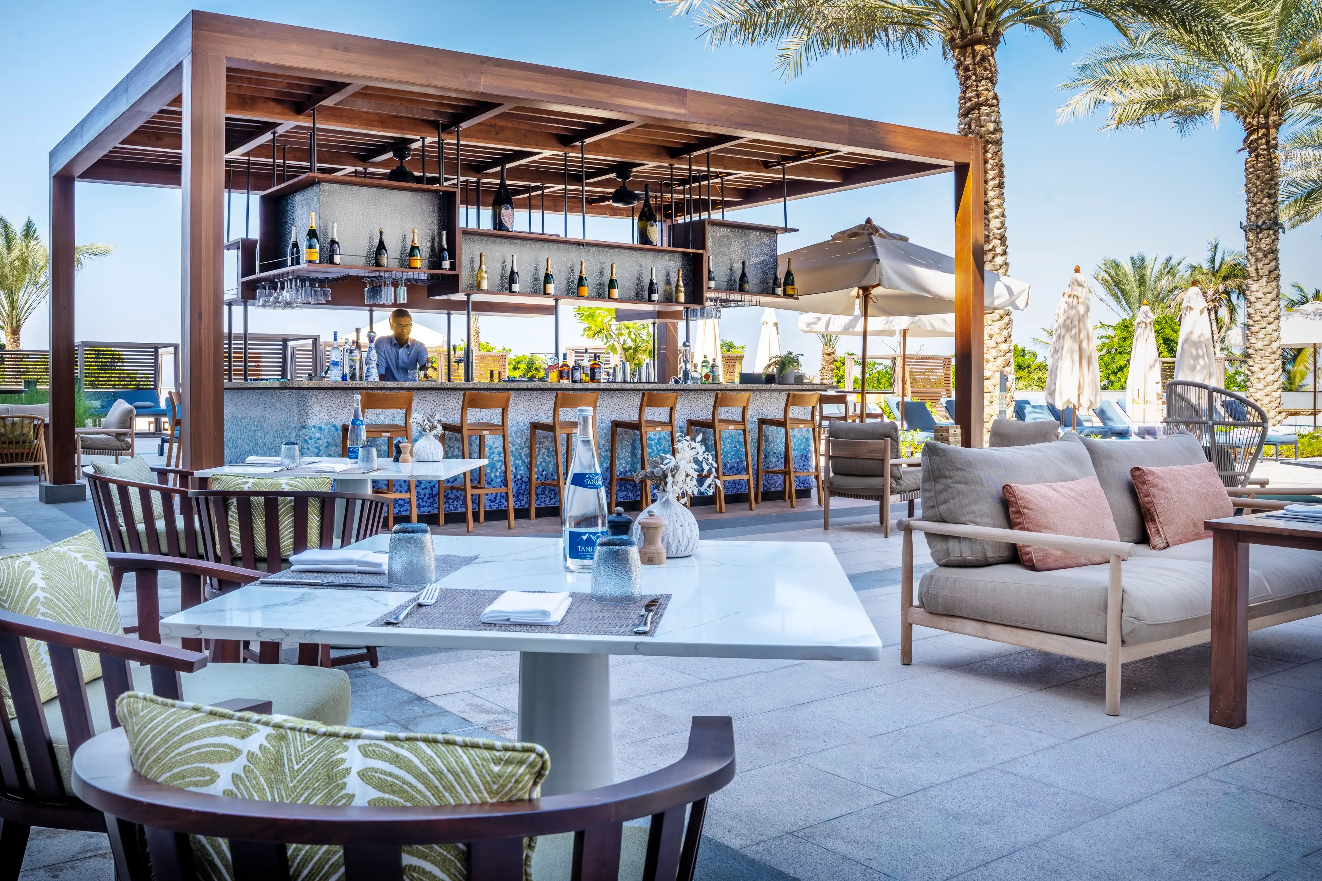 Timber-framed bar counter with stools, lounge sofas and palm trees under a clear sky at The Pool Bar, Mandarin Oriental, Muscat