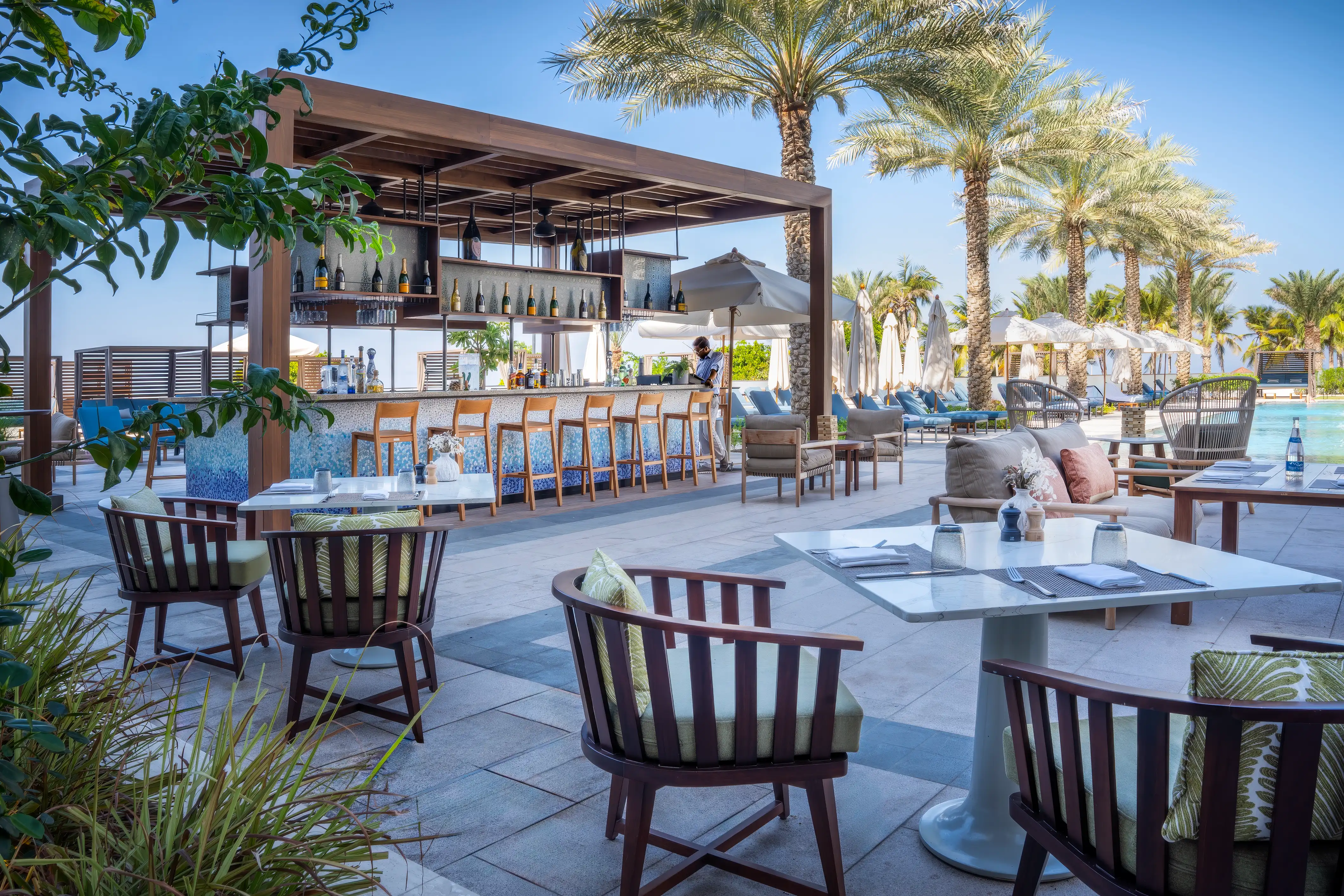 Outdoor terrace seating beside the pergola-covered bar, with sun loungers, the pool and palm trees beyond, at The Pool Bar, Mandarin Oriental, Muscat