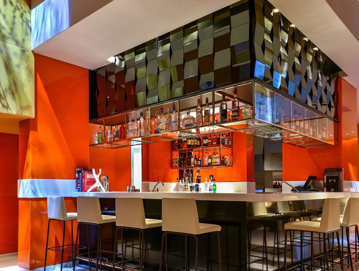 The Living Room bar at W Amman with orange lacquered walls, white bar stools and a faceted mirrored canopy over the bottle display
