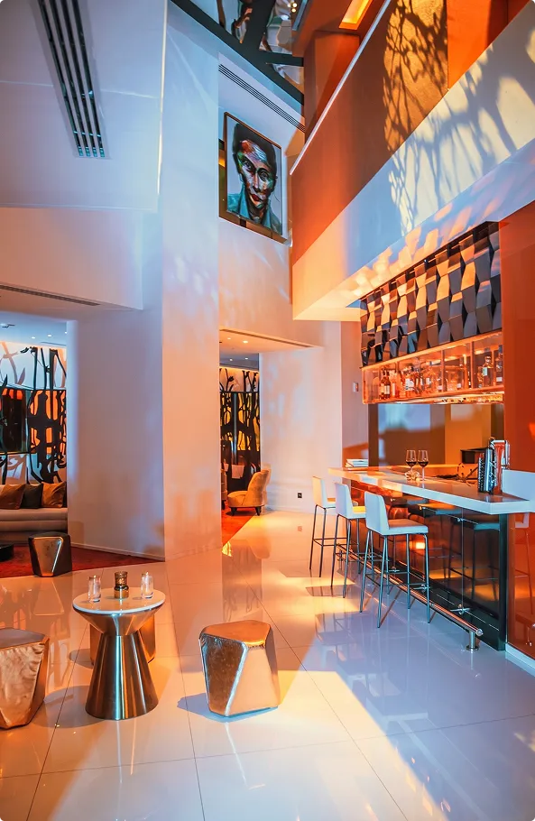 The Living Room at W Amman with metallic side tables, a white bar counter, a portrait artwork and projected leaf patterns on the walls