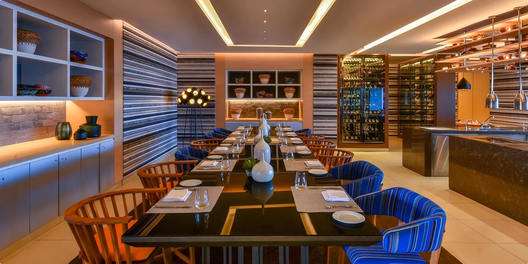 Mesh dining area at W Amman with a long communal table, striped blue upholstered chairs and a glass fronted wine display