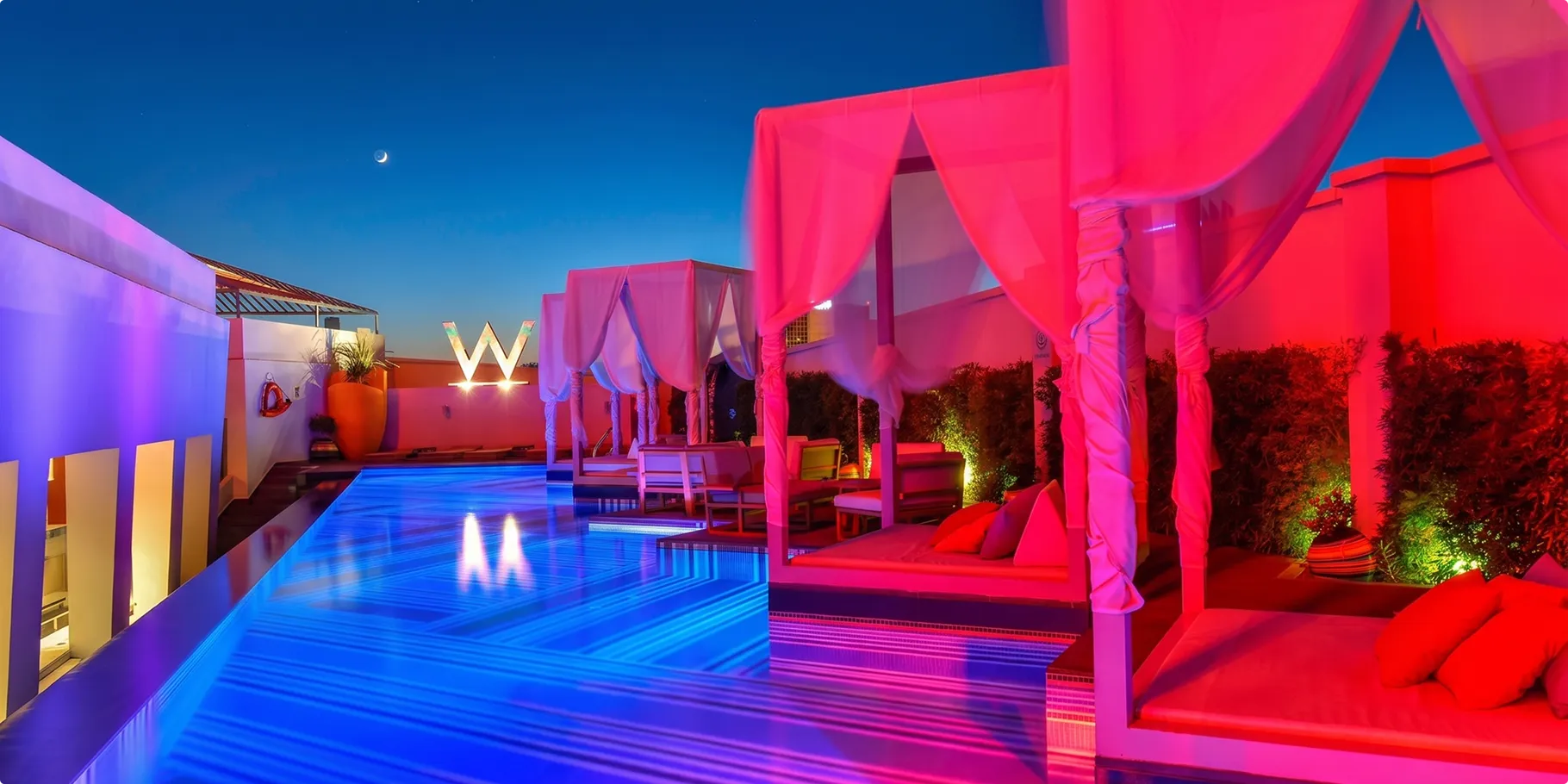 WET Deck pool at W Amman at dusk, lit in blue and pink, with curtained cabanas and the W sign