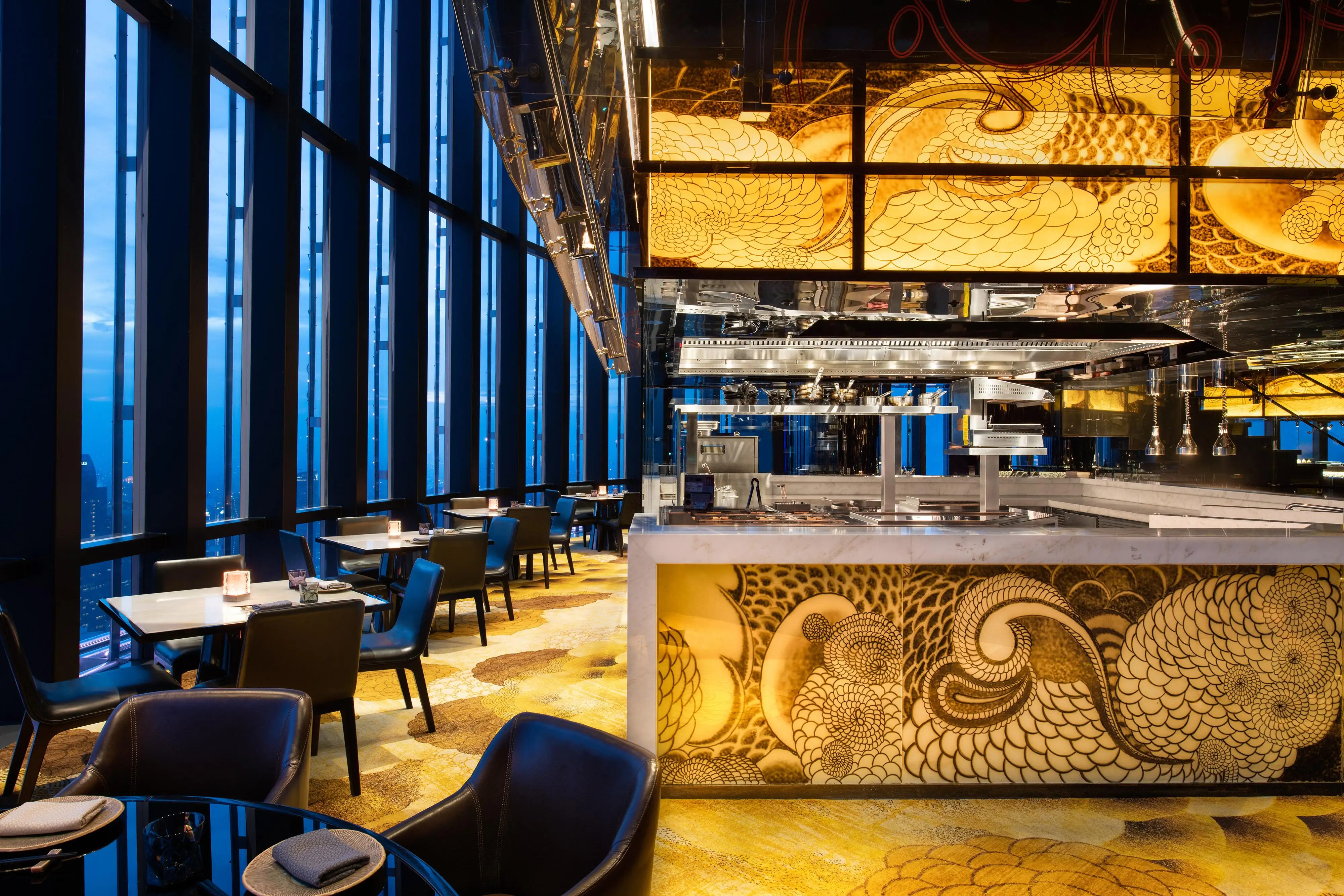 Open kitchen at Henshin, The Westin Jakarta, with a swirl-patterned counter front, backlit amber panels and window-side dining tables