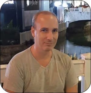 Man in a light gray shirt sitting outdoors near a water canal at night.
