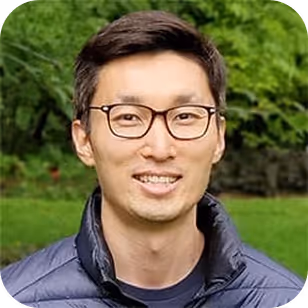 Smiling man with glasses and short hair wearing a dark jacket outdoors with green foliage background.