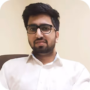 Man with dark hair, beard, and glasses wearing a white shirt, seated against a beige background.