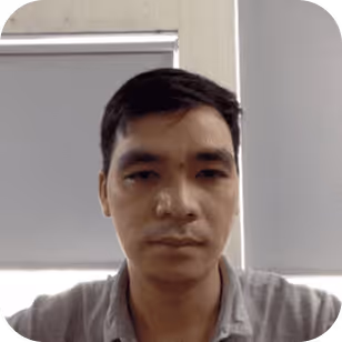 Portrait of a man with short black hair wearing a gray shirt in front of a window with closed blinds.