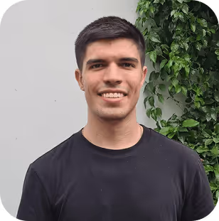 Young man with short dark hair wearing a black shirt smiling against a gray wall with green foliage background.