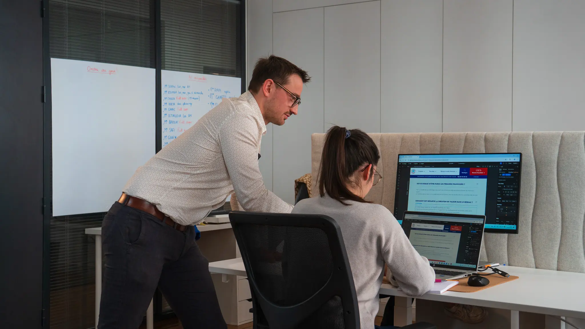 Un homme debout aidant une femme assise à un bureau devant deux écrans d'ordinateur dans un bureau moderne.