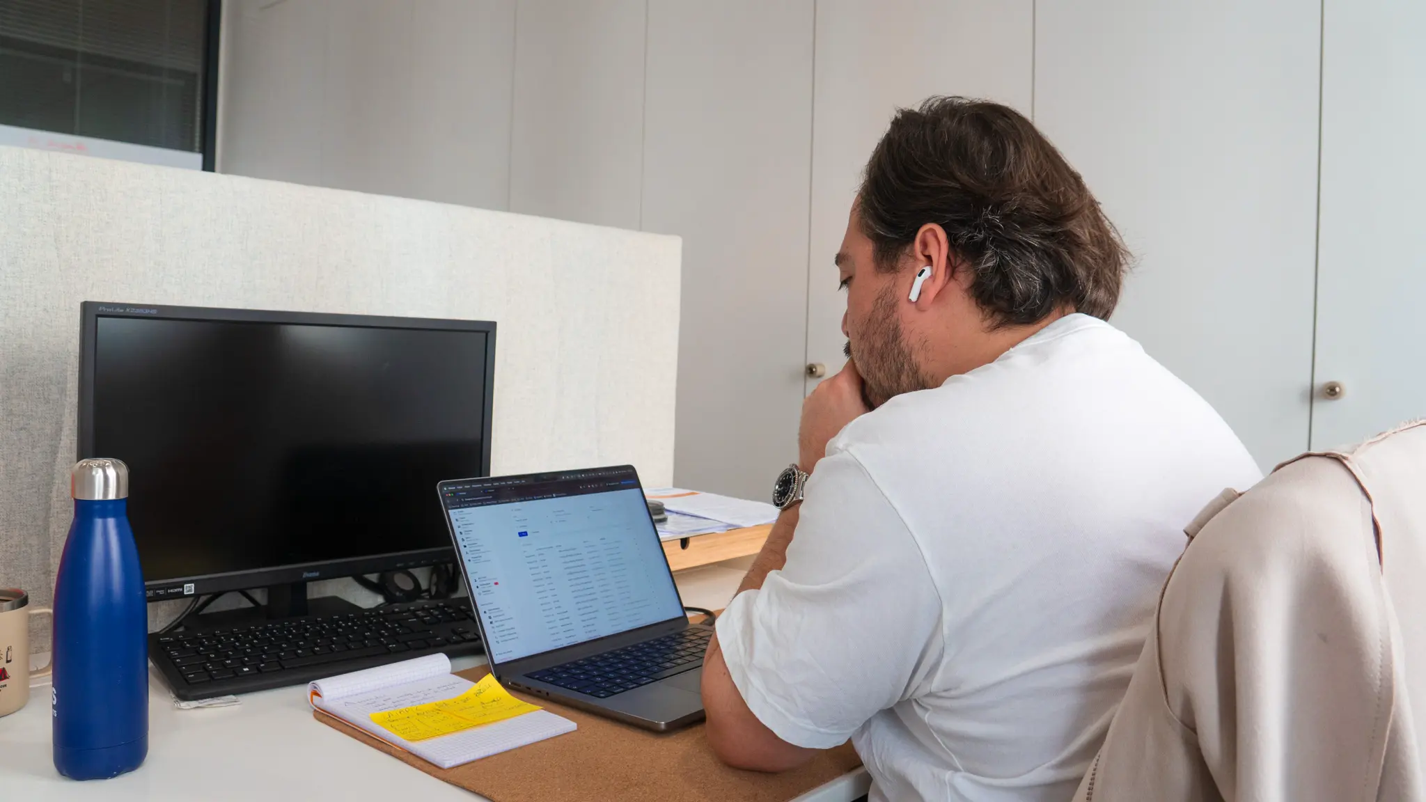 Un homme portant un t-shirt blanc et des écouteurs sans fil travaille sur un ordinateur portable à son bureau avec un écran d'ordinateur éteint, une bouteille bleue et des notes manuscrites à côté.