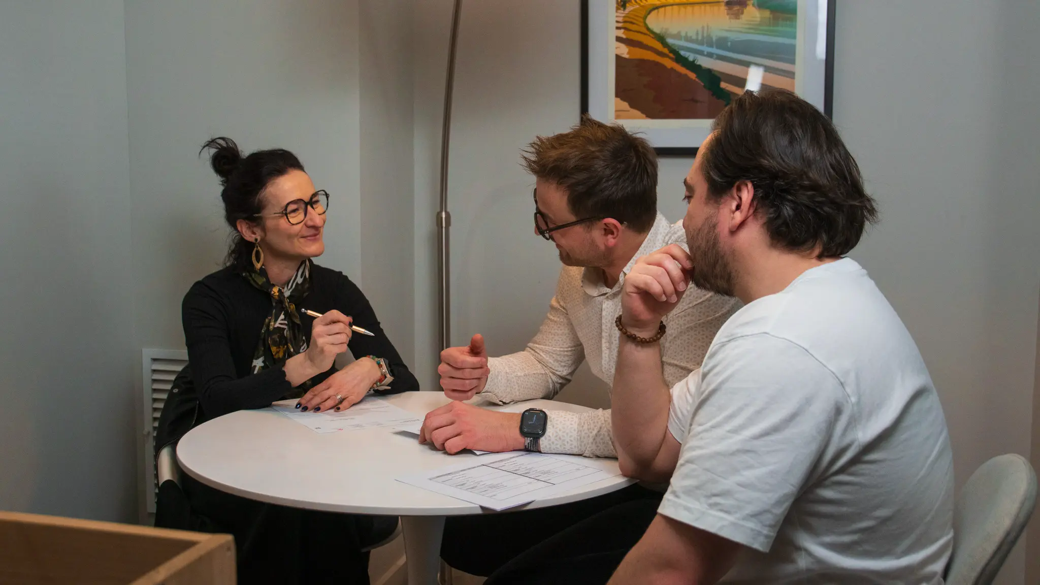 Trois personnes discutent ensemble autour d'une table ronde blanche dans un bureau, avec des documents devant eux.