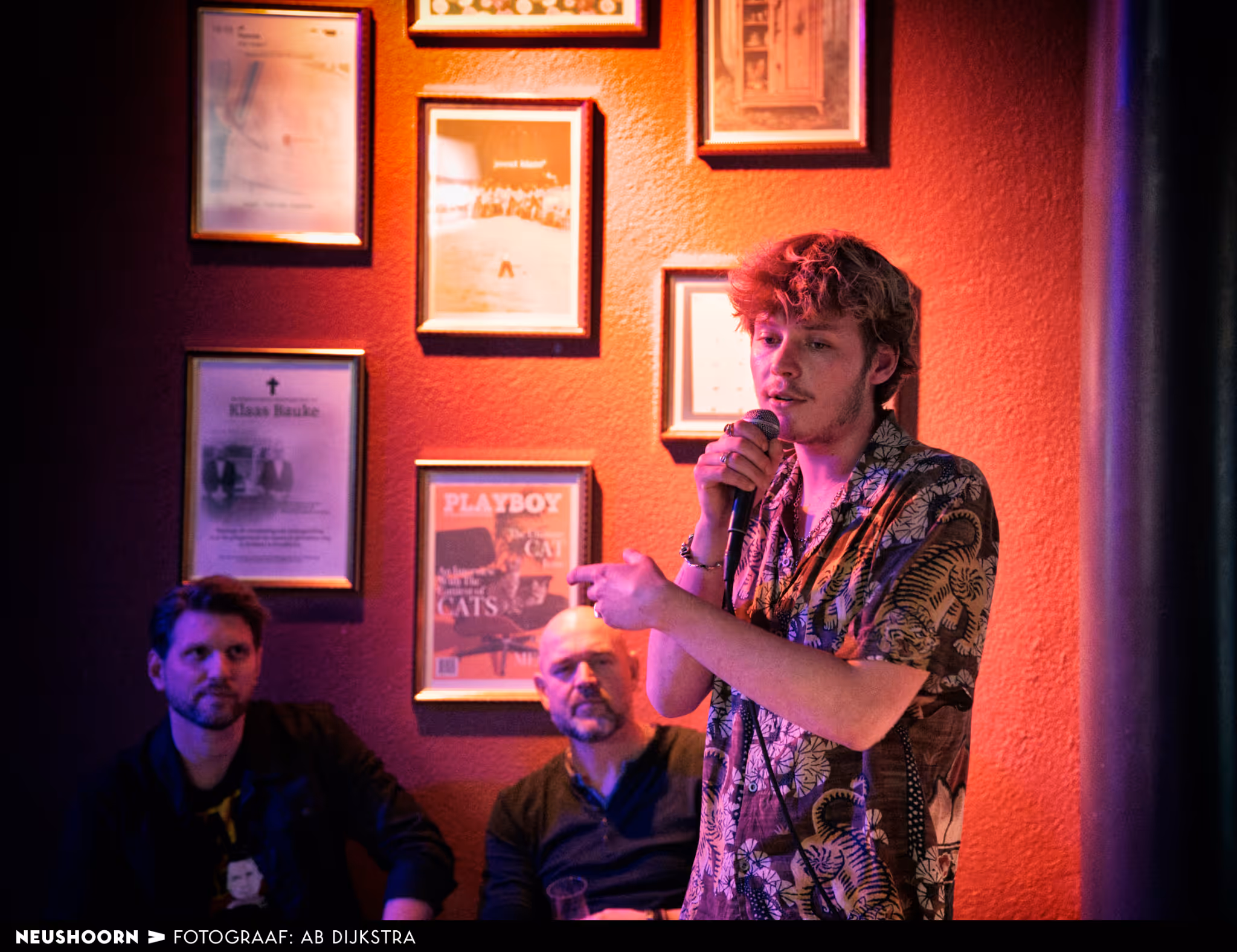 Een jongeman houdt een toespraak voor mensen in de Bunker Bar, op de achtergrond kijken twee mannen aandacht toe. Er hangen gouden lijsten op de muur achter hen.