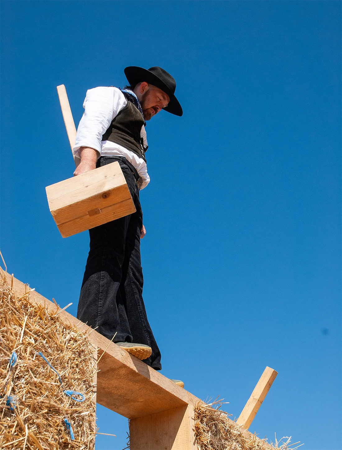 Zimmermann in traditioneller Kluft und schwarzem Hut balanciert mit Holzklotz auf einer Strohbalken-Konstruktion vor strahlend blauem Himmel.