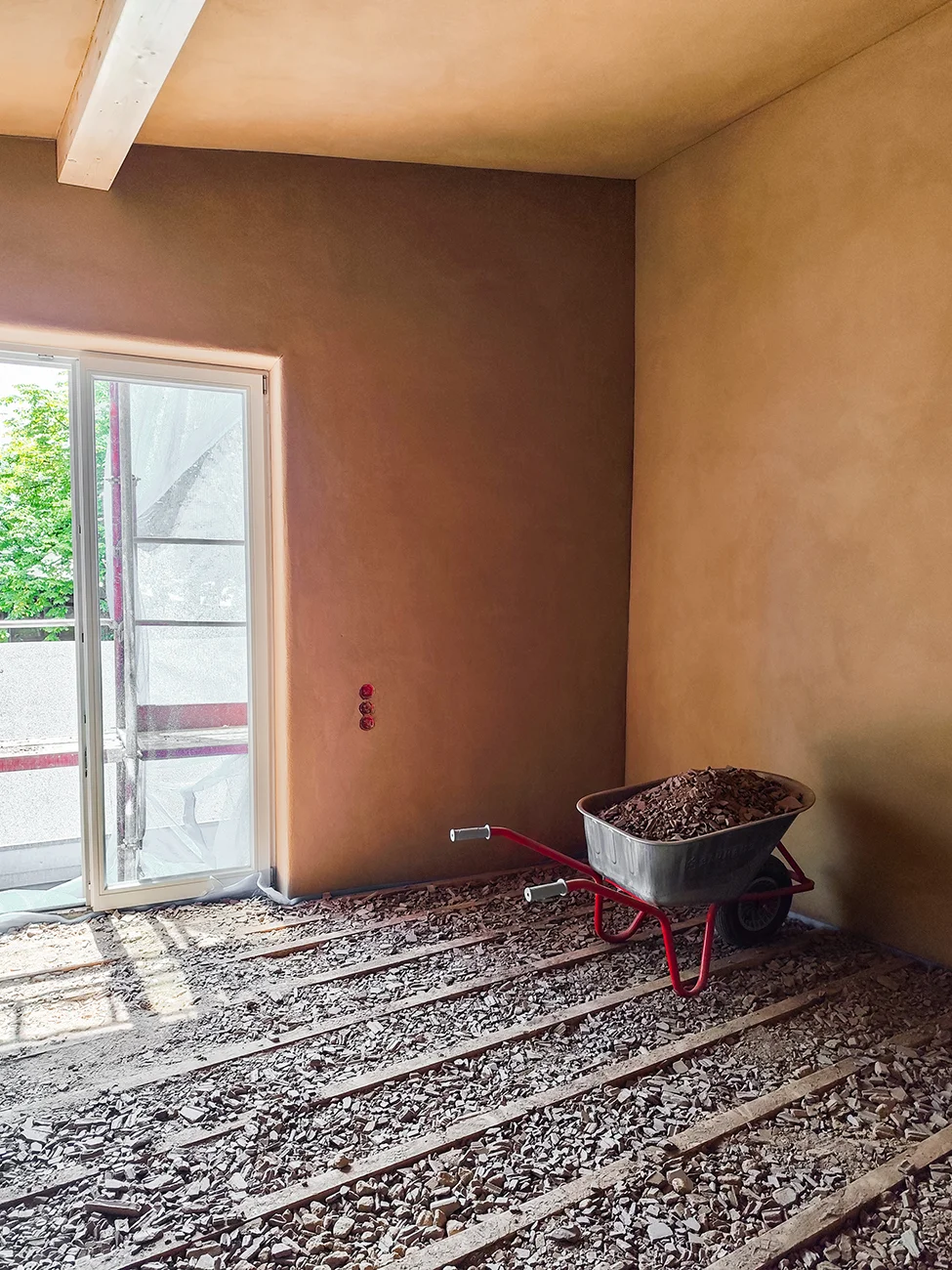 Lehmverputztes Zimmer im Ausbau: Schubkarre mit Schotterboden vor offenem Fenster, Projekt von Takatukabau.