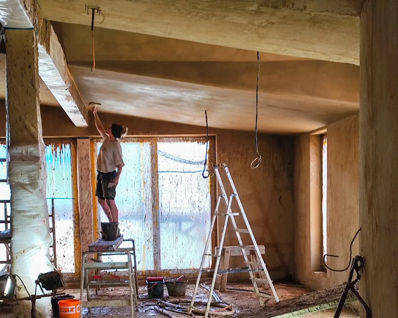 Handwerker glättet Lehmputz an Decke; Leiter und Werkzeug in naturbelassenem Rohbau, Sanierungsprojekt von Takatukabau.