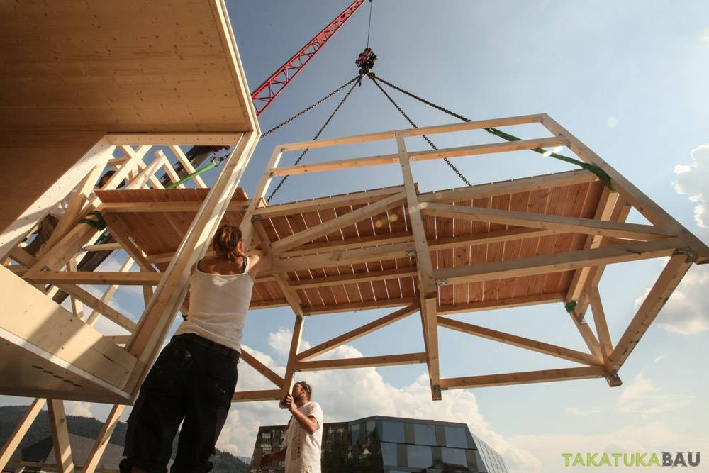 Kran setzt Holzmodul auf Dach; zwei Zimmerer von Takatukabau koordinieren die Montage hoch über Freiburgs Innenstadt.
