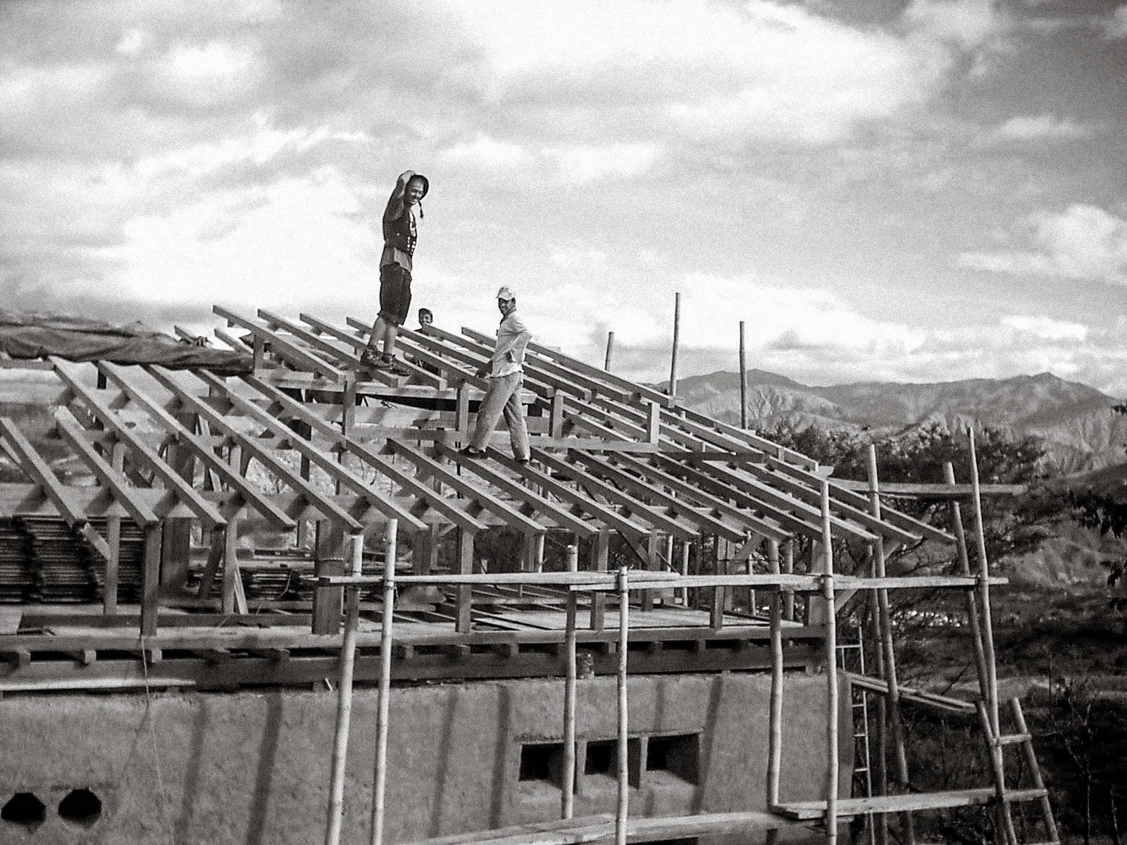 Zimmerer steht auf dem Holz-Dachstuhl eines frühen Bauprojekts; das Gebäude aus Lehm befindet sich noch in der Rohbauphase – Gerüste und Berge im Hintergrund.