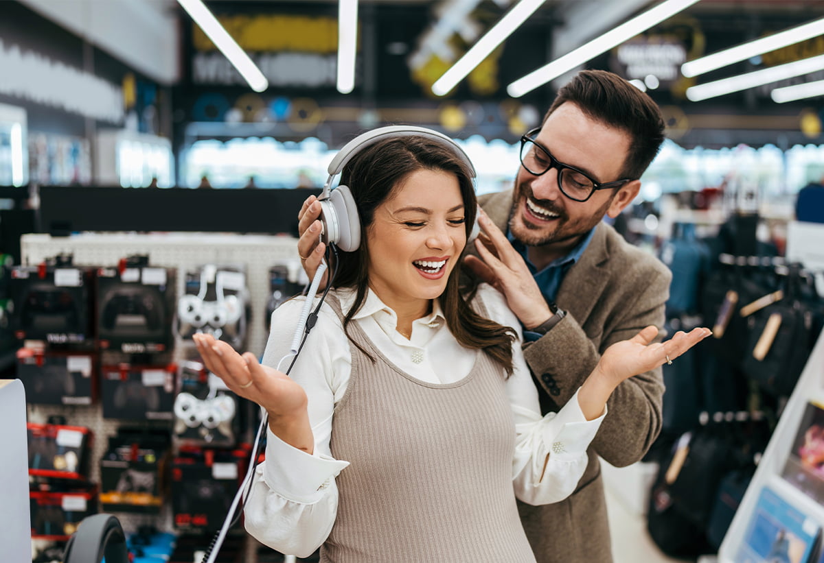 Zwei Personen testen gemeinsam Kopfhörer in einem Elektronik-Store, die Kundin trägt Over-Ear-Kopfhörer und reagiert begeistert.