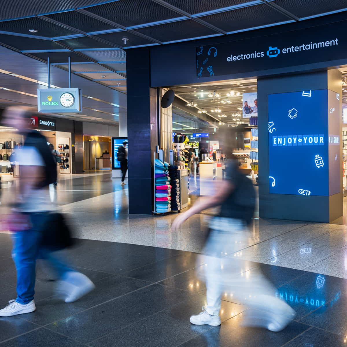 Elektronik- und Entertainment-Shop am Flughafen Frankfurt, vorbeigehende Reisende mit Bewegungsunschärfe im Vordergrund, beleuchtete Verkaufsfläche mit Zubehör und Digitalanzeigen im Hintergrund.