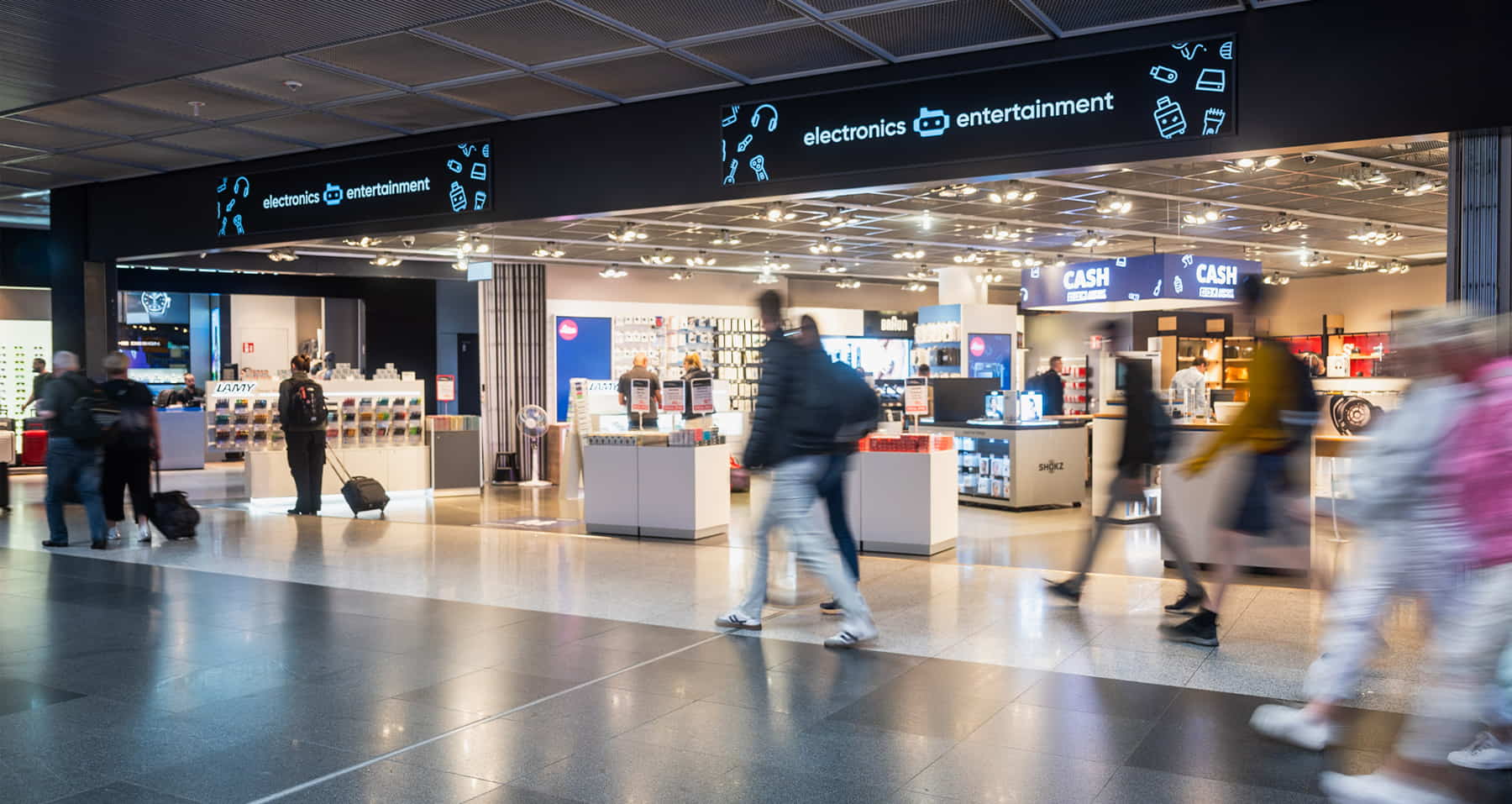 Elektronik- und Entertainment-Shop von PAX Airport Shopping in einem Flughafen-Terminal, helle Verkaufsfläche mit Produktinseln, vorbeigehende Reisende mit Bewegungsunschärfe im Vordergrund.