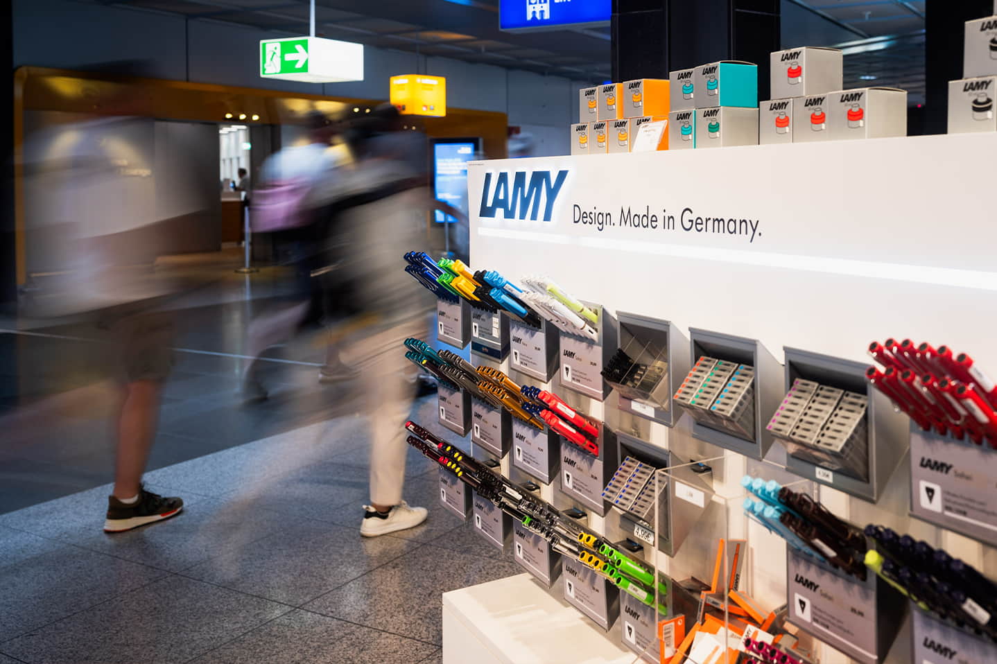 LAMY-Display mit Schreibgeräten im Flughafen-Einzelhandel, übersichtliche Produktpräsentation, Reisende passieren den Shopbereich.
