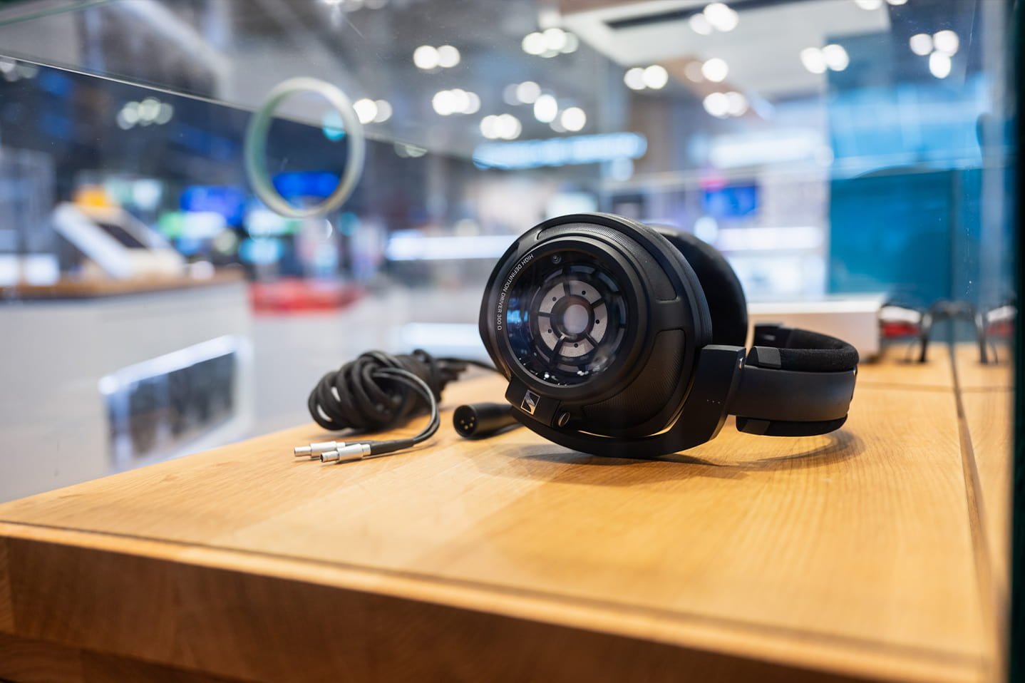 Over-Ear-Kopfhörer mit Kabel auf einer Holzpräsentationsfläche in einem Elektronikgeschäft, unscharfer Verkaufsbereich im Hintergrund.