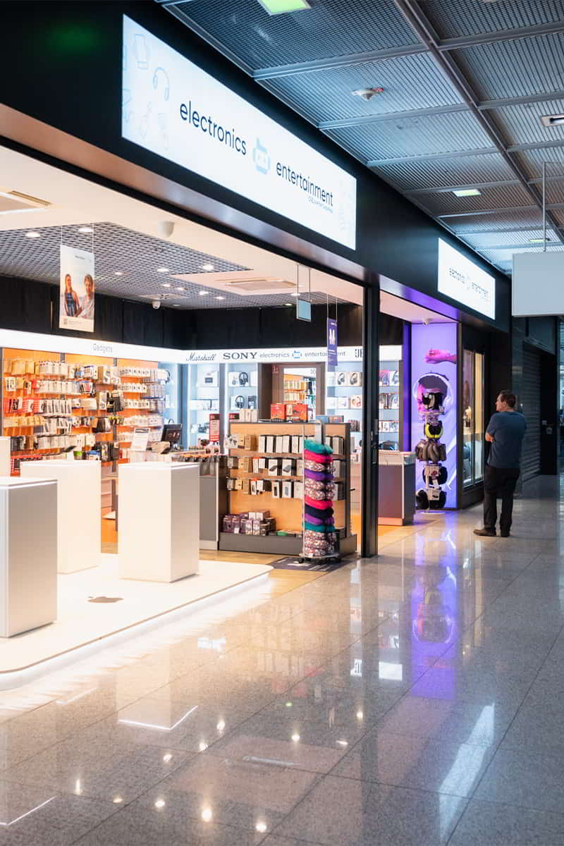 Elektronik- und Entertainment-Shop von PAX Airport Shopping im Flughafen-Terminal, offene Verkaufsfläche mit Technikzubehör und Markenprodukten, beleuchtete Shopfront entlang des Gangs.