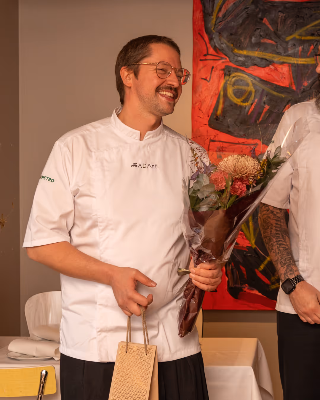 Marco hymyilee valkoisessa esiliinassa, kädessään kukkakimppu ja lahjapussi.