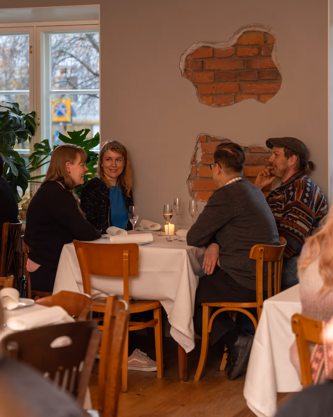Neljä ihmistä istuu pöydän ympärillä ravintolassa keskustellen, taustalla sisäseinän tiiliseinä paljastuneena.